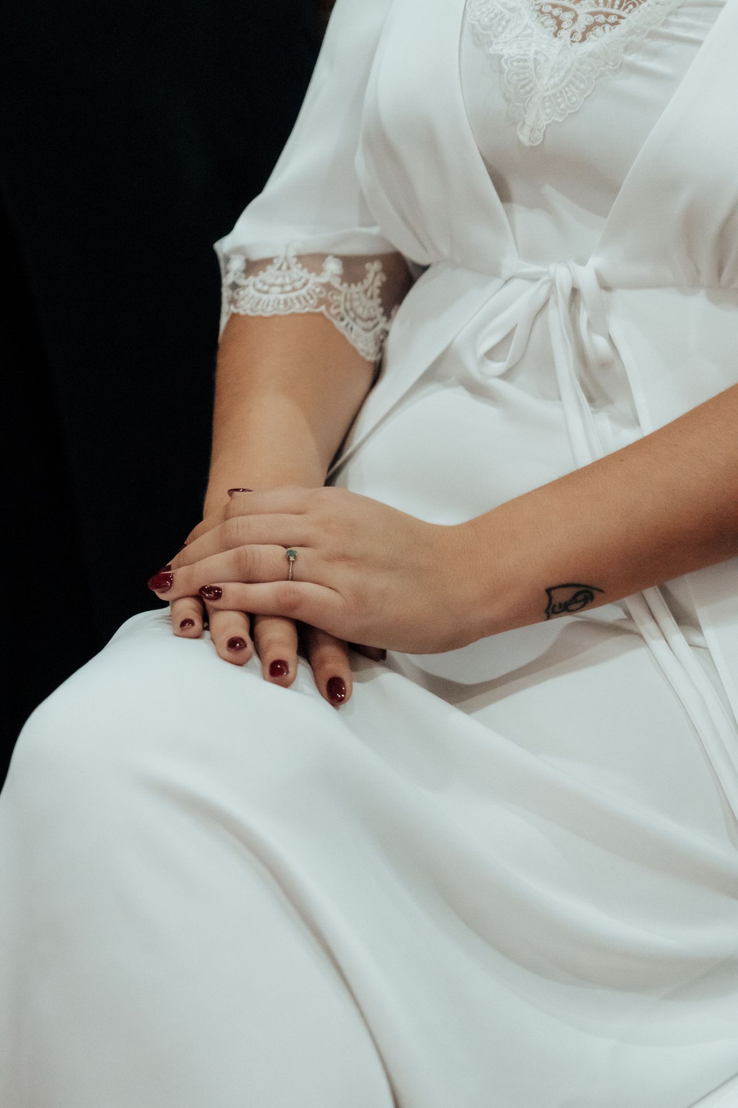 Close-up of a bride's hands with dark red nails and engagement ring resting on white satin robe.