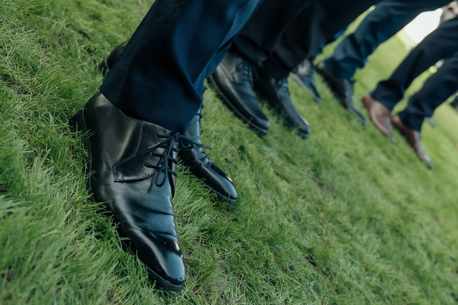 Close-up of groomsmen wearing polished black dress shoes and navy suits standing on lush green grass at a wedding.