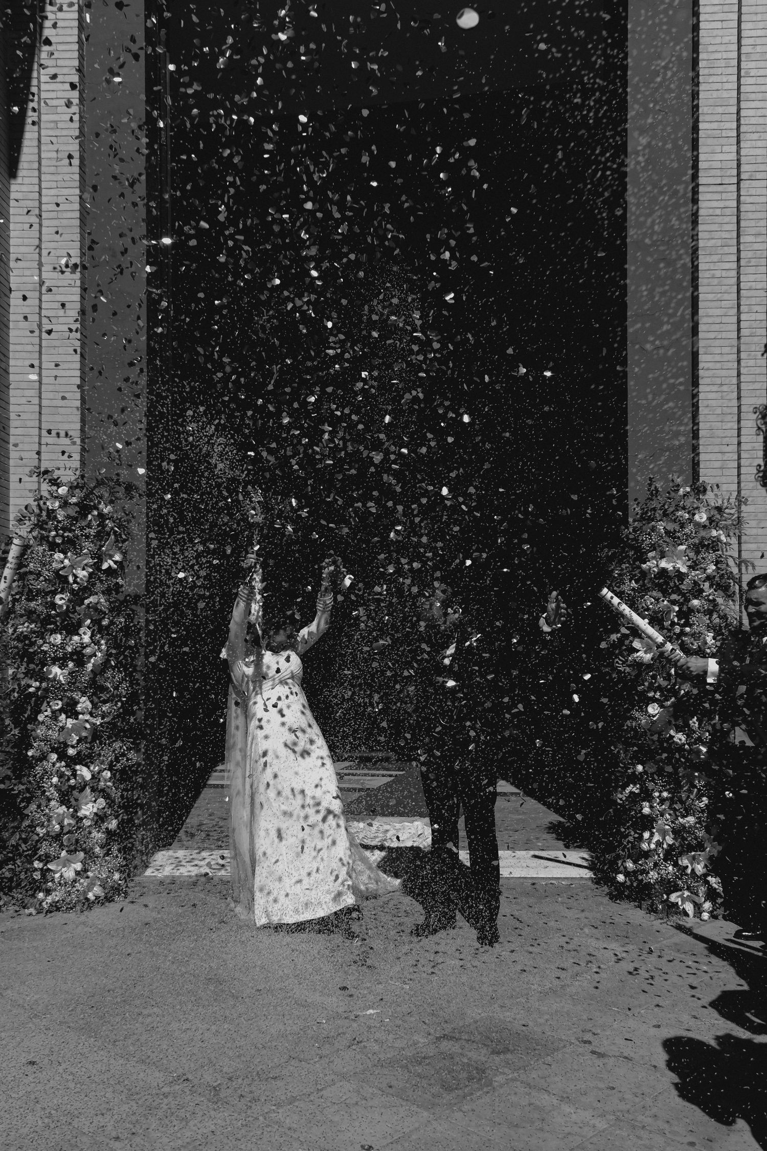 Black and white photo of newlyweds surrounded by confetti shower outside church doors.