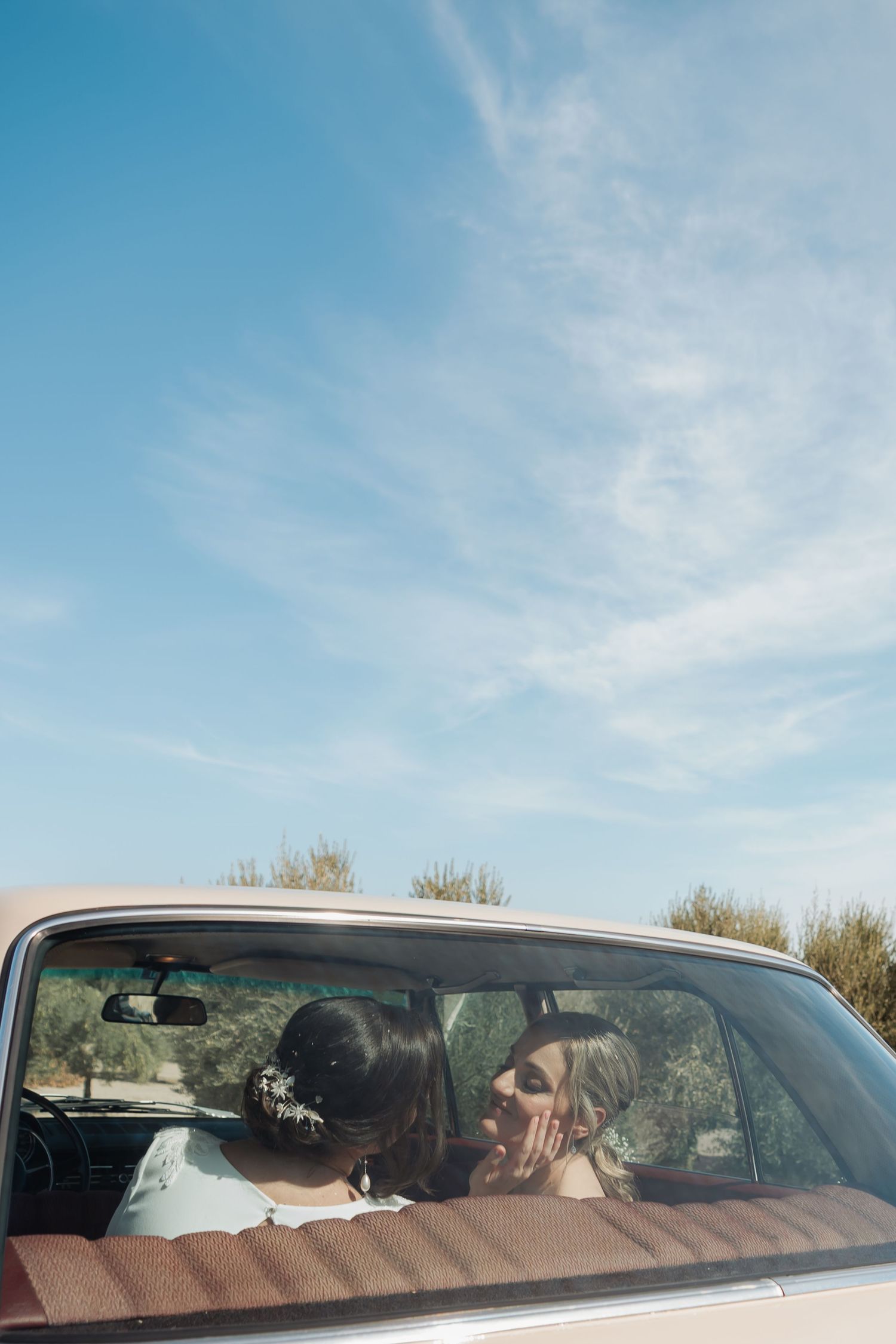 Two brides share a tender moment inside a vintage car under a bright blue sky.