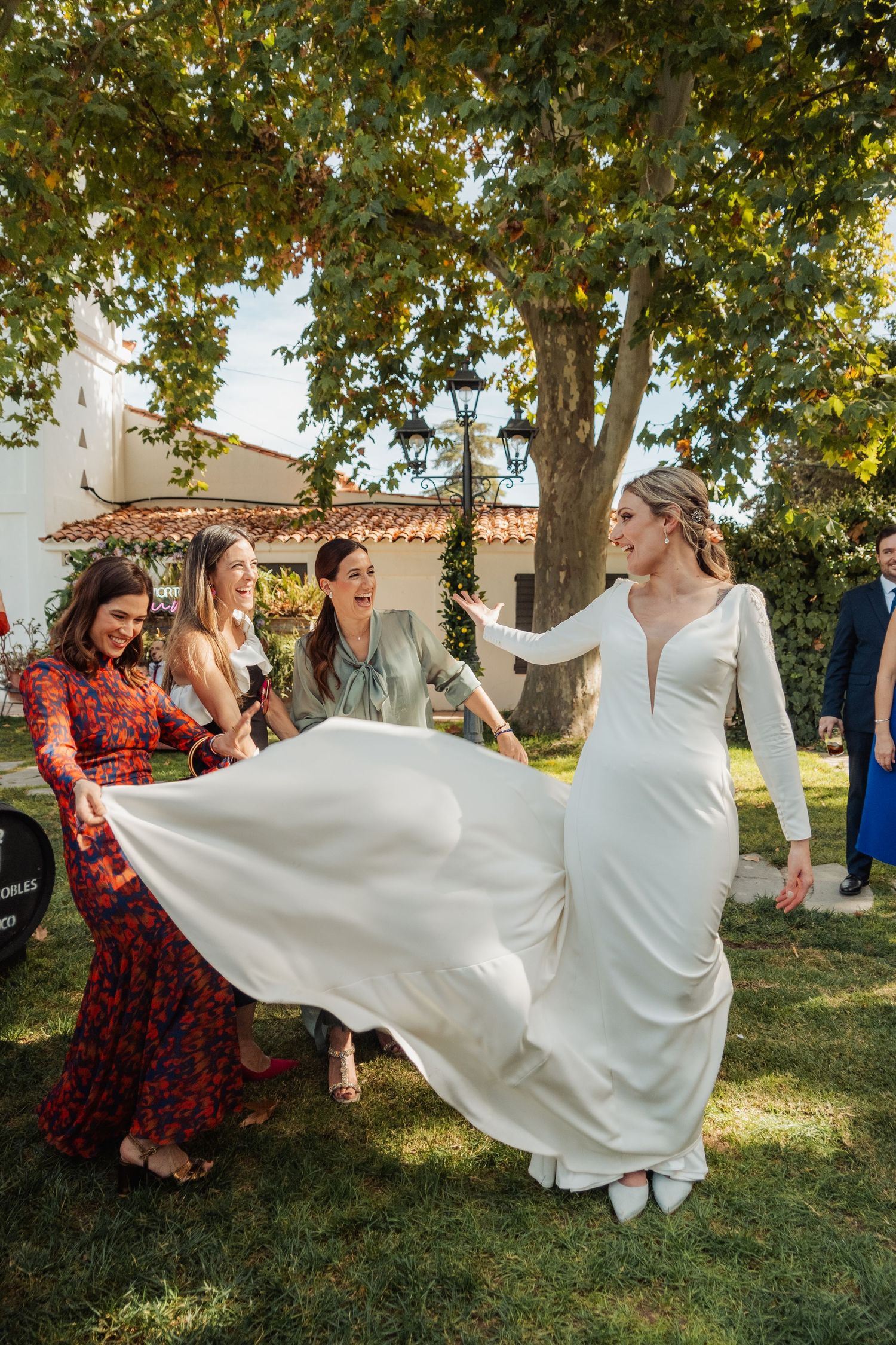 Bride in white gown laughs with friends as they playfully lift her dress outdoors.