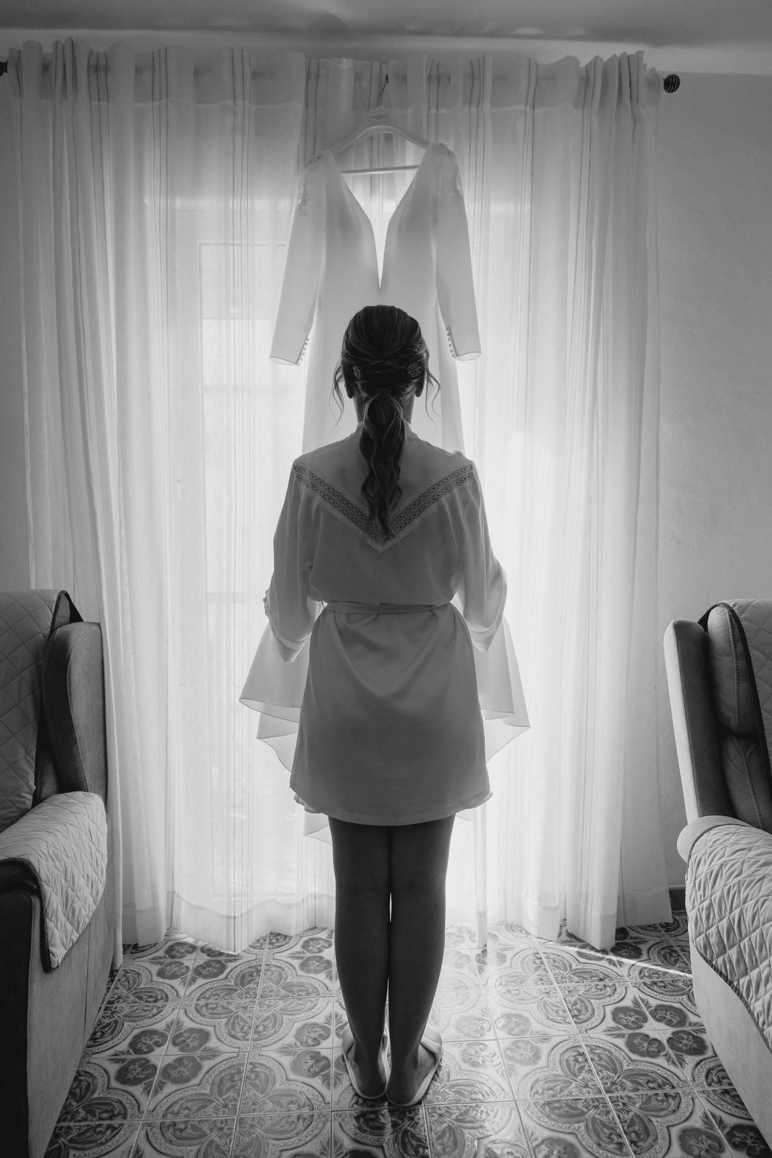 Black and white photo of bride in robe standing before hanging wedding dress at window.