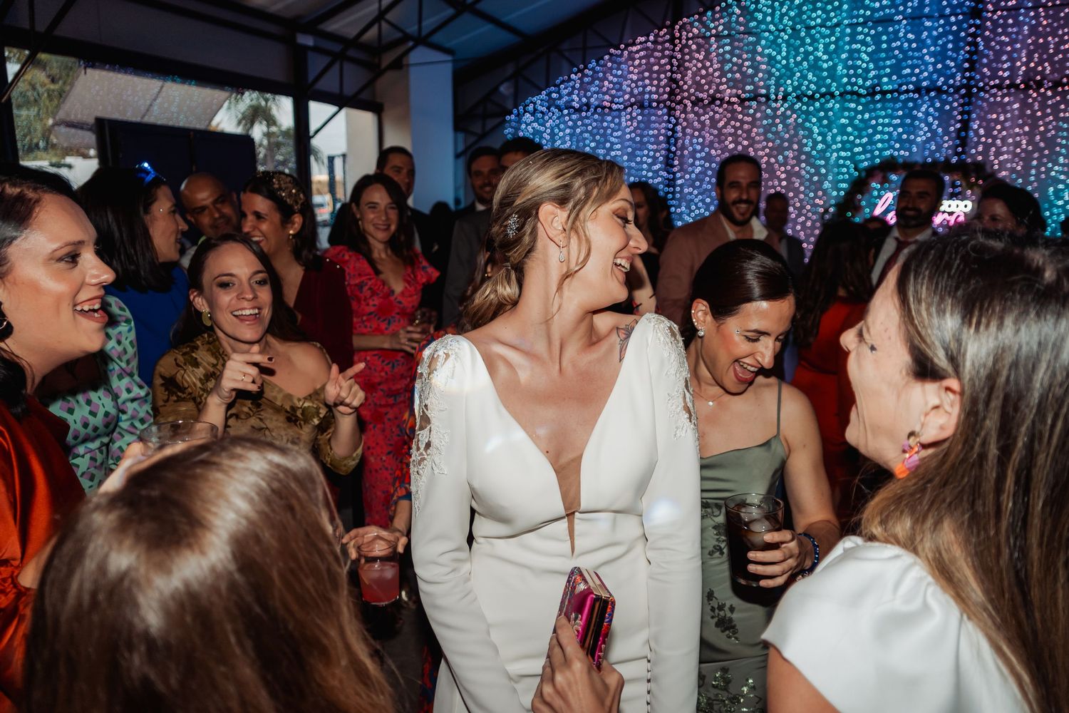 Blonde bride in deep V-neck white gown smiling and mingling with guests at a vibrant wedding reception.