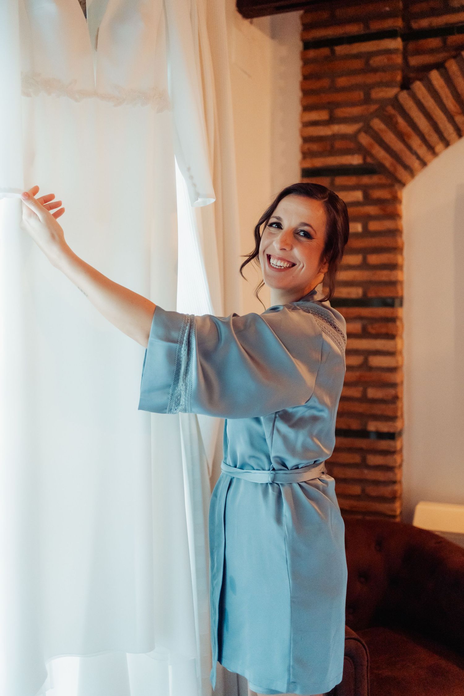 Smiling bride in blue satin robe holding white wedding dress near brick wall window.