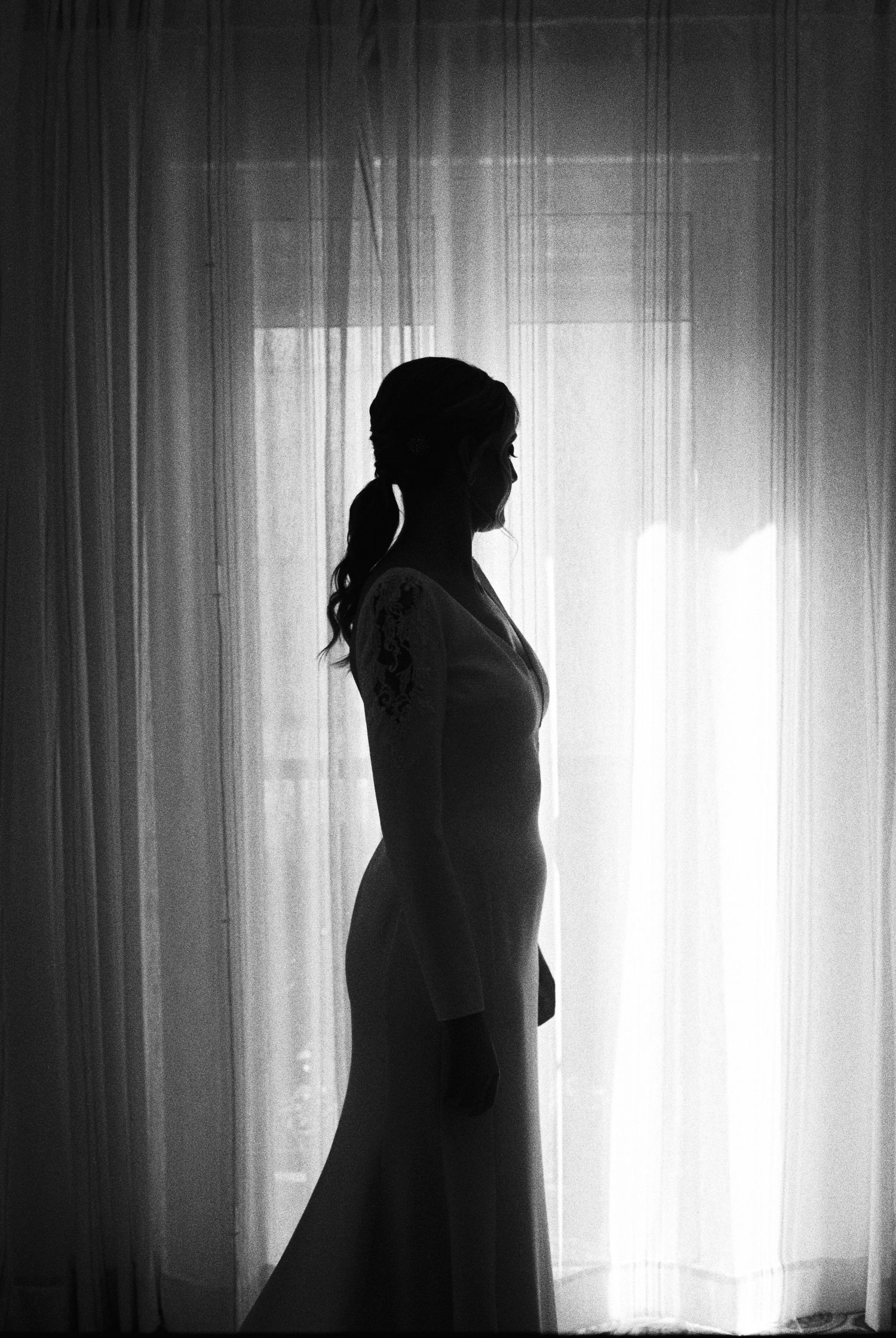 Elegant black and white silhouette of bride in wedding gown standing before sheer curtained window.