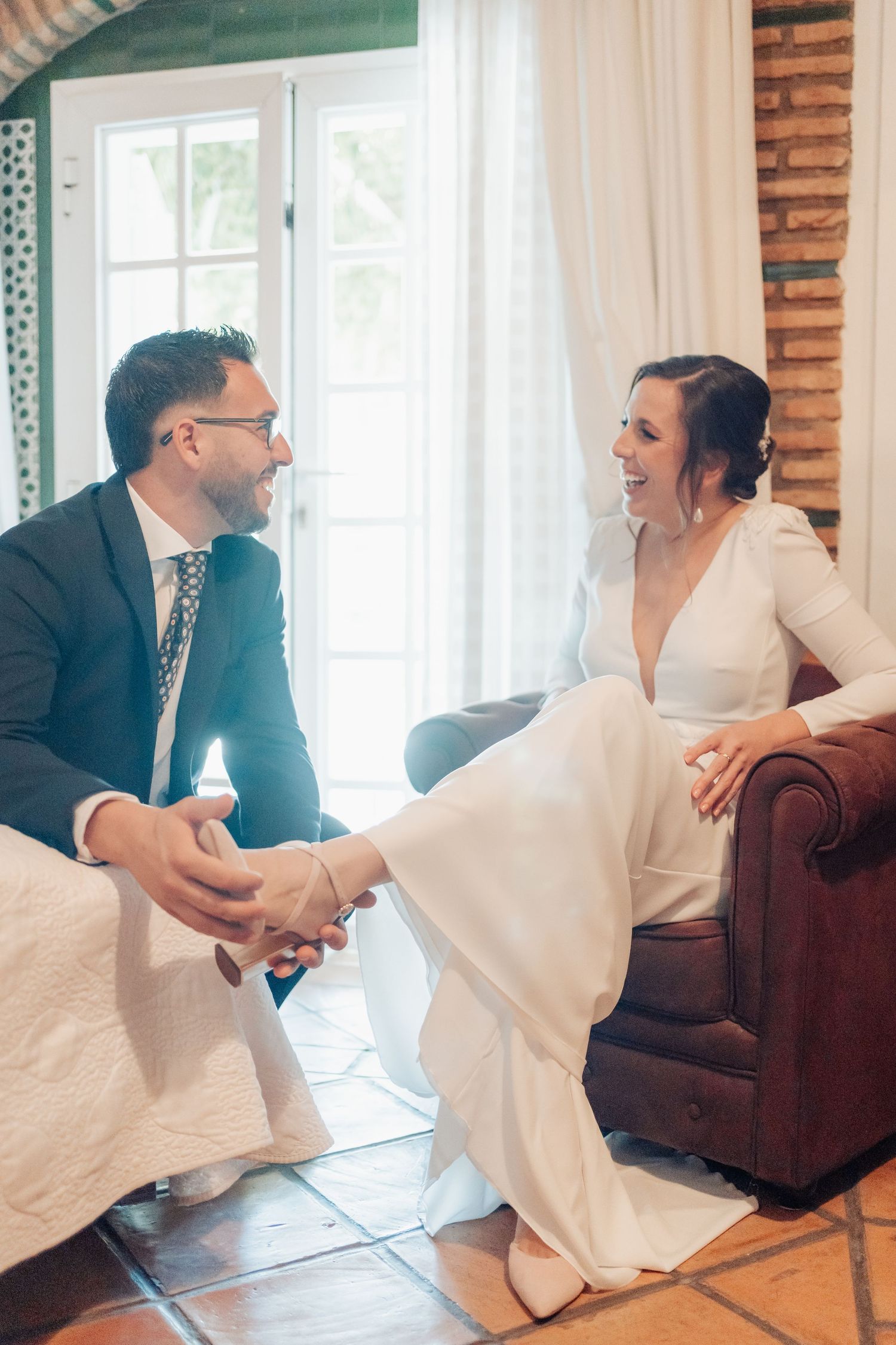 Laughing bride in white gown and groom in suit share relaxed moment together indoors.