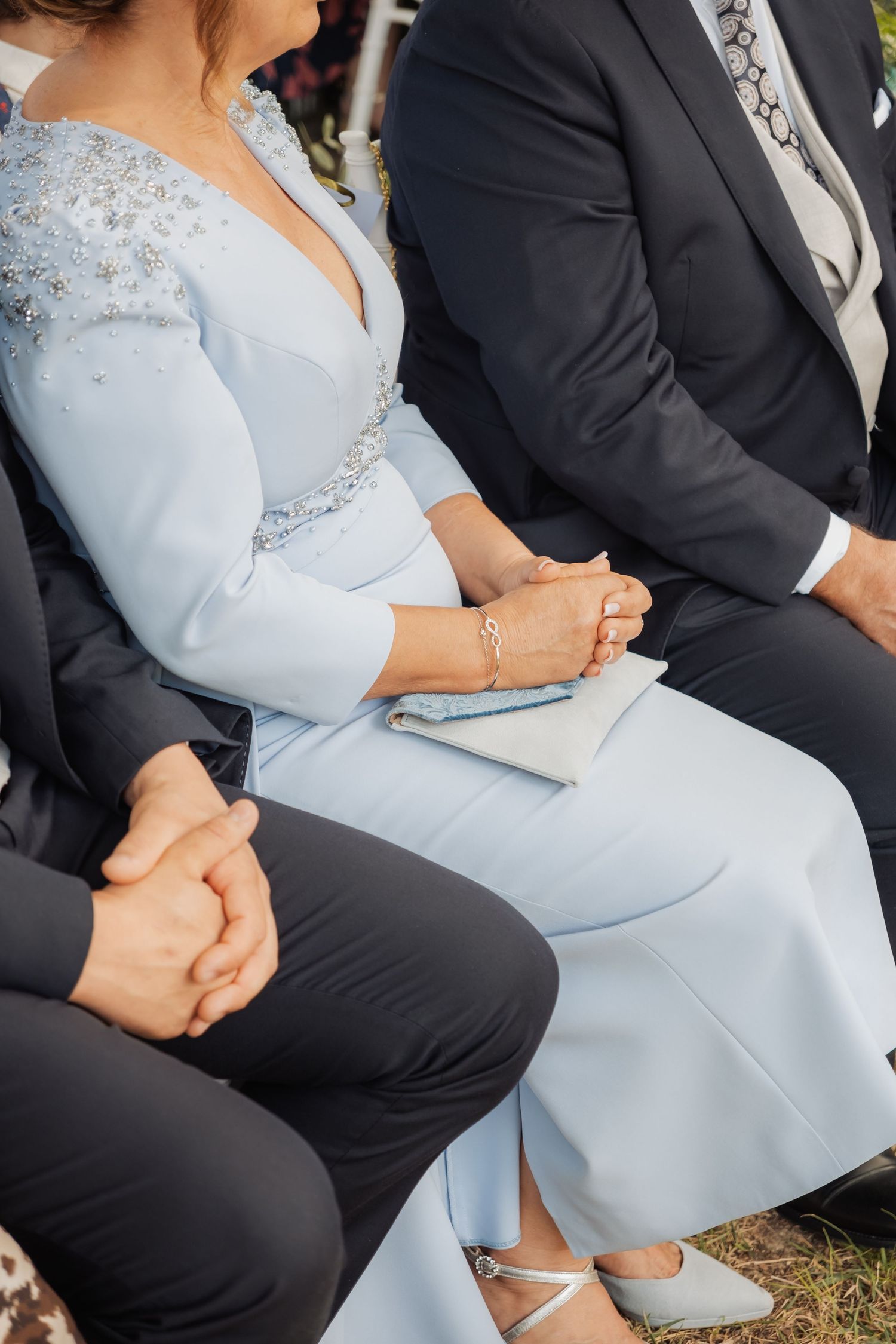 Elegantly dressed wedding guests in formal attire sit together during an outdoor wedding ceremony on grass.