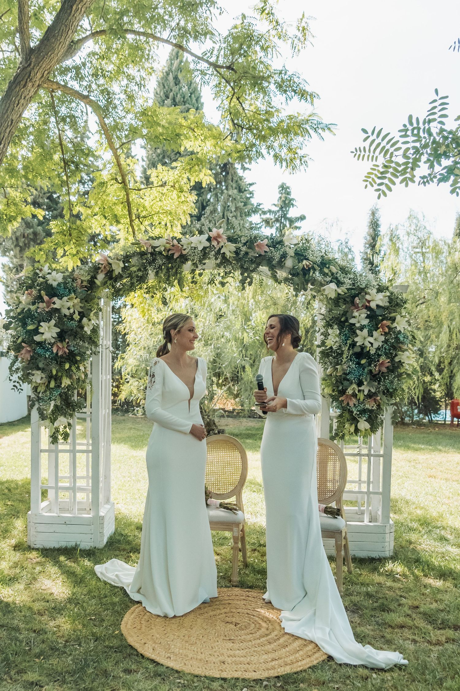 Two brides in white gowns laughing together under a floral arch at an outdoor garden wedding.