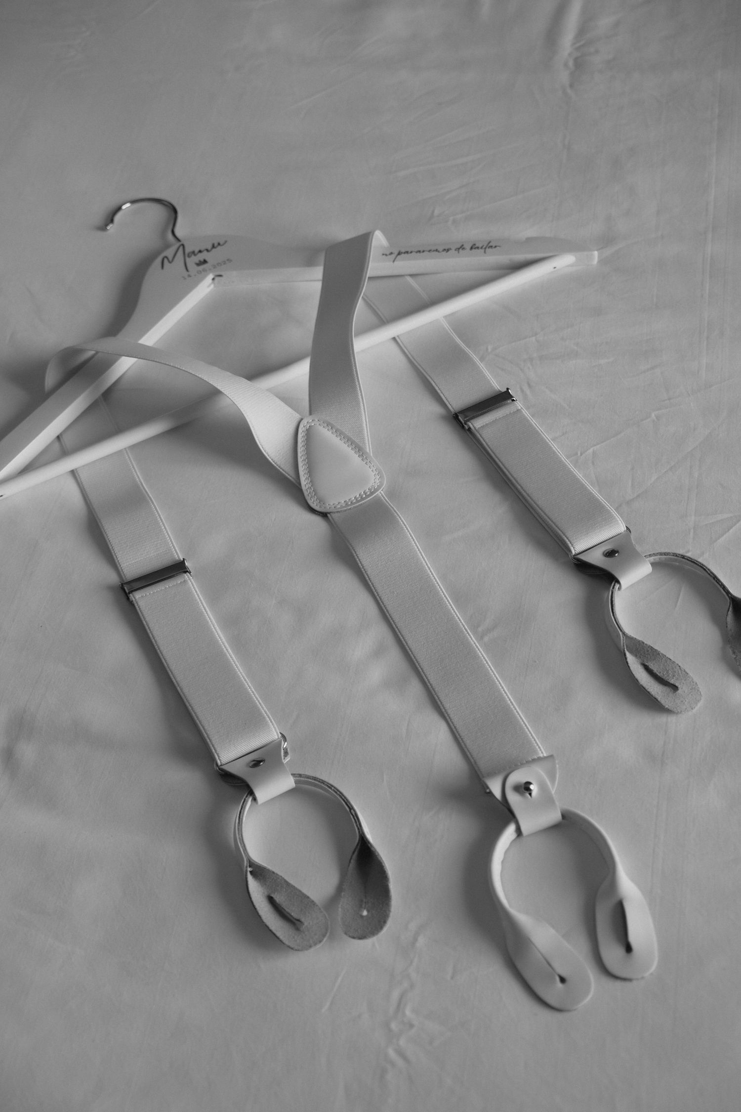 Black and white flat lay of white suspenders and a personalized wedding hanger on a white surface.