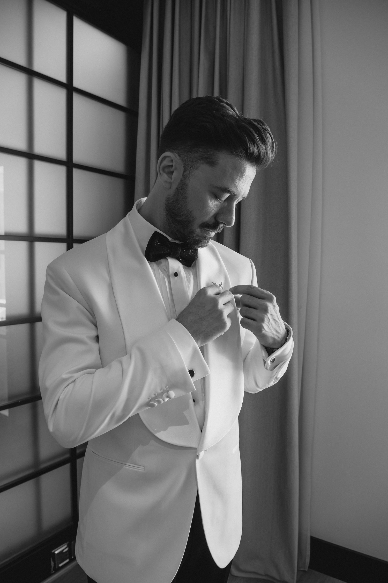Black and white portrait of a groom in white tuxedo pinning a boutonniere near a window.