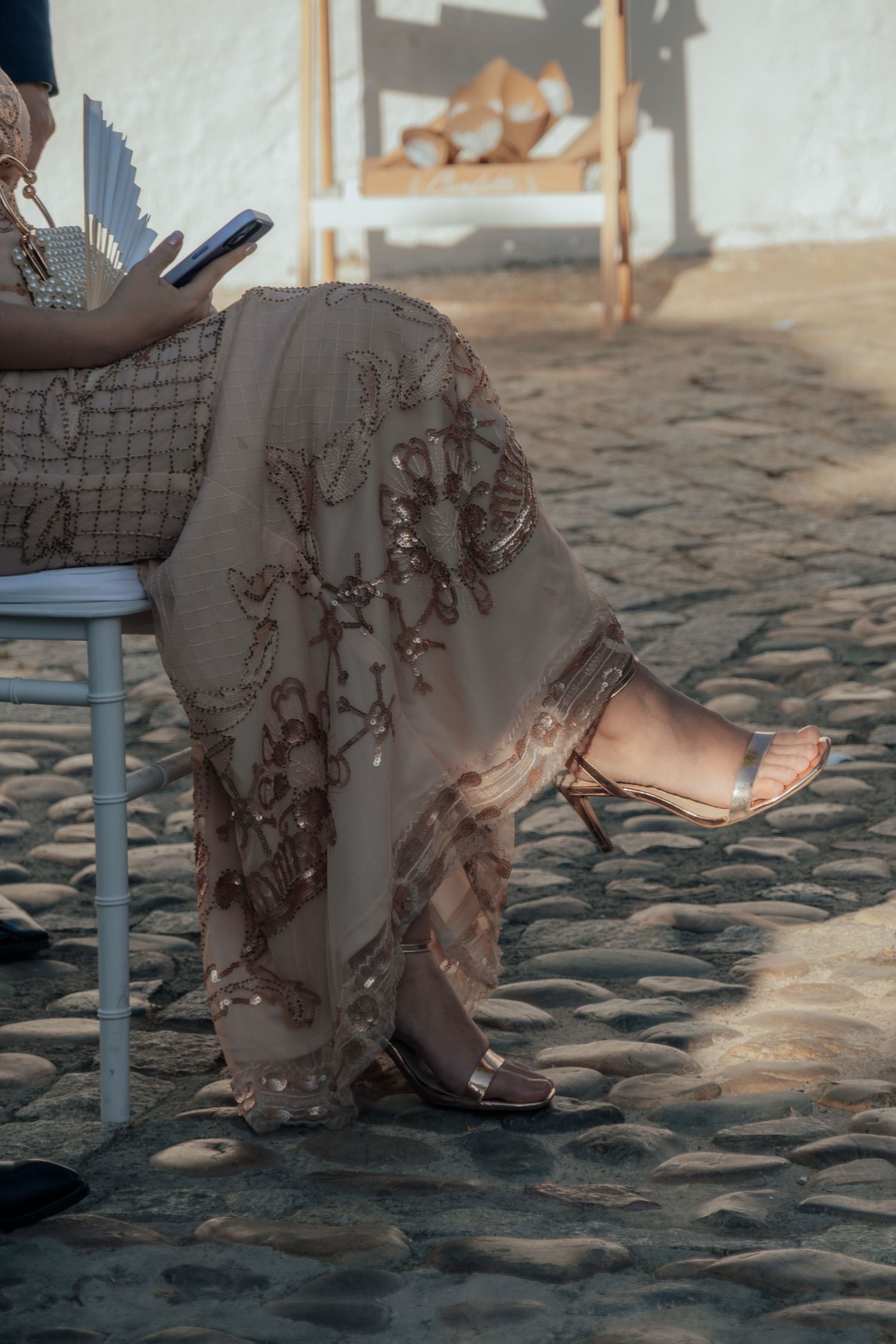 Woman in embroidered beige gown seated on chair at outdoor wedding, holding phone on cobblestone.