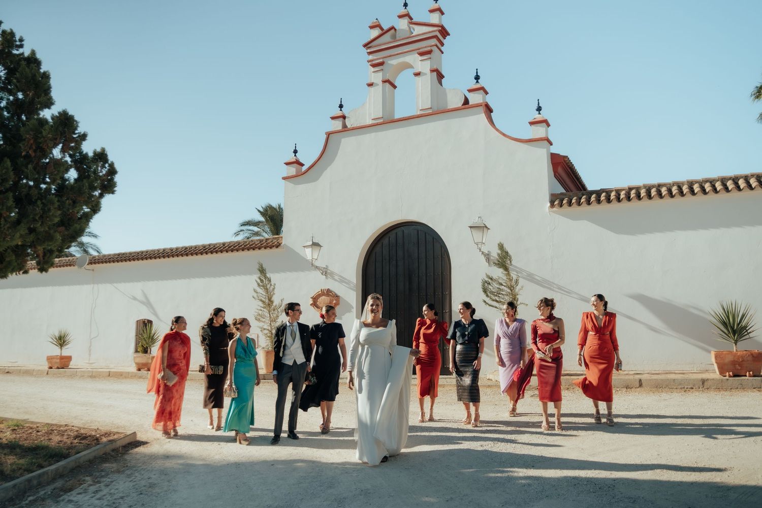 Bride and wedding party walking together in front of a white Spanish colonial chapel in sunlight.