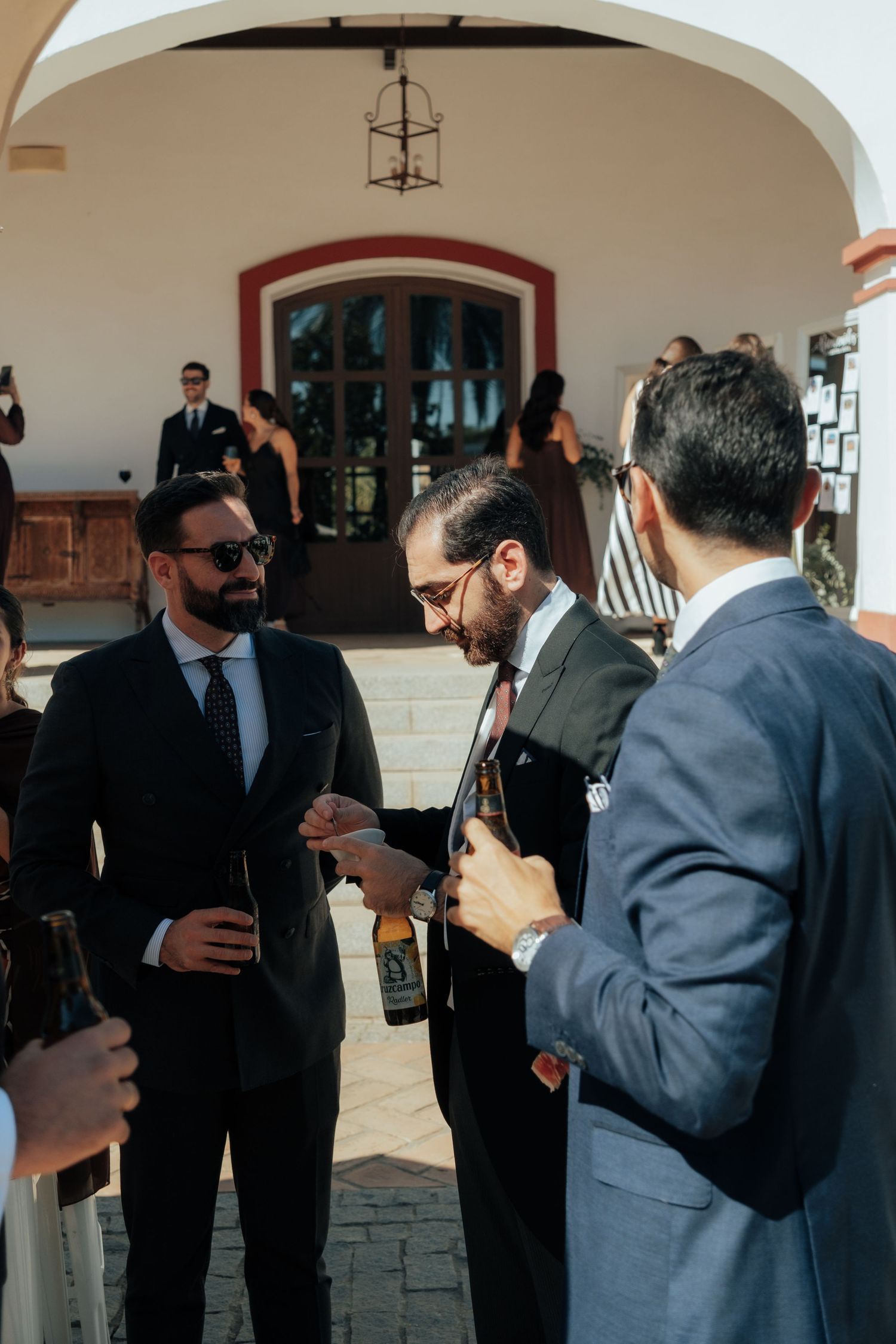 Three well-dressed men in suits drinking beer outside a white wedding venue.