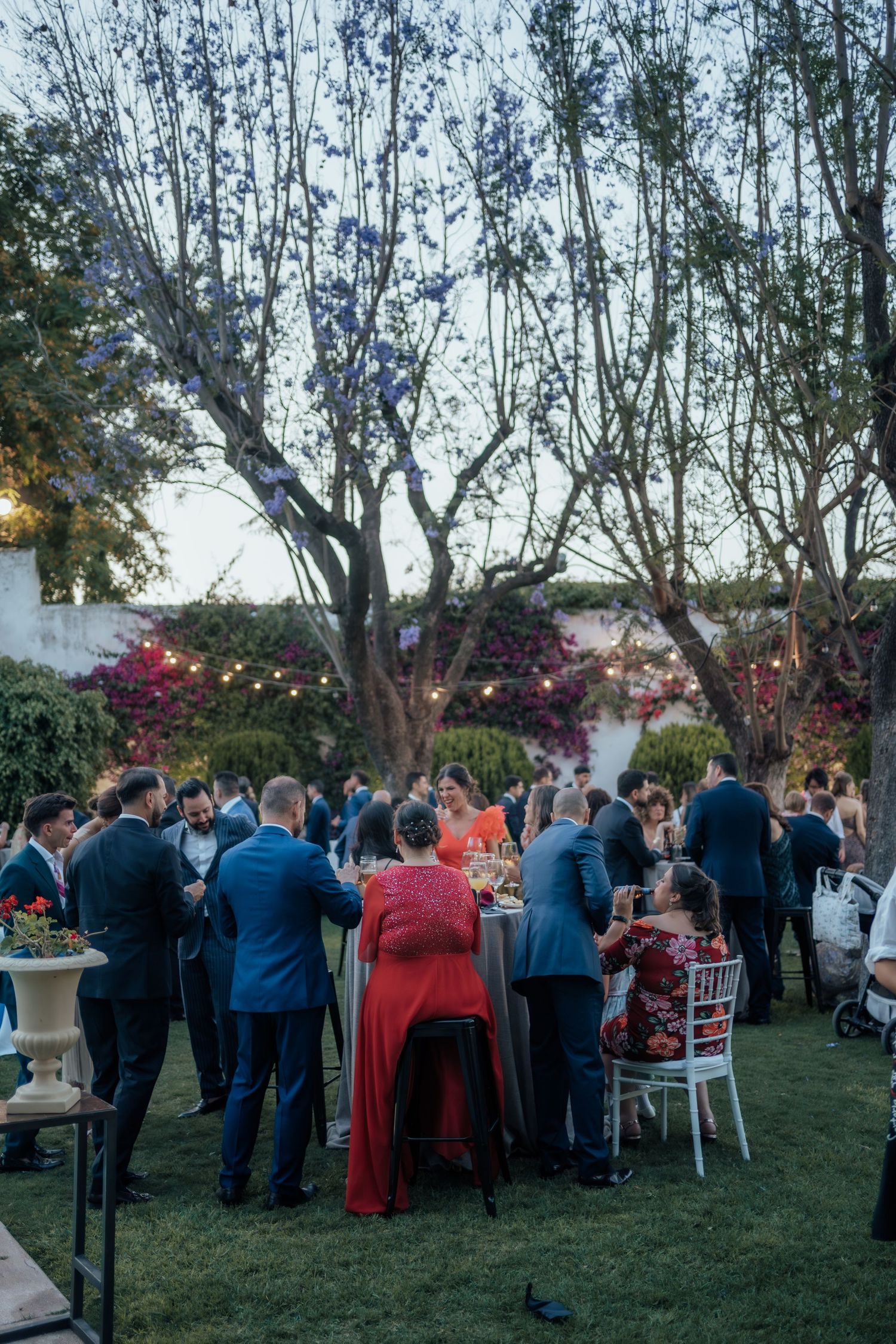Wedding guests dining at decorated outdoor garden reception tables at dusk.