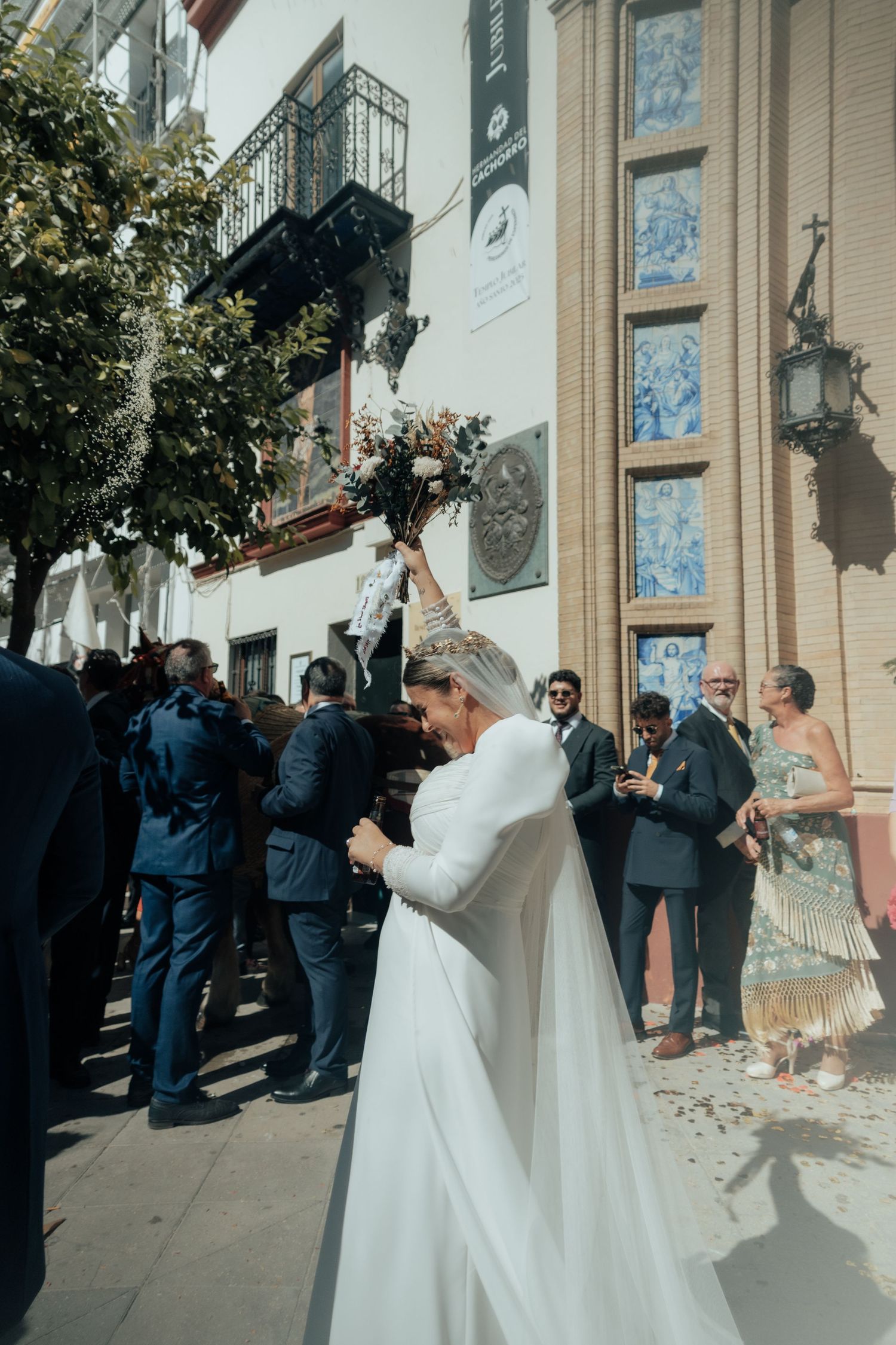 Bride in white gown raises bouquet joyfully outside a decorated Spanish church amid wedding guests.