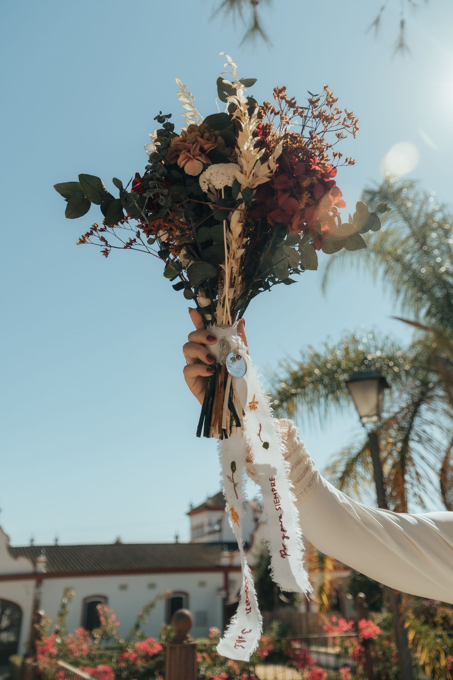 Bride triumphantly raises rustic bridal bouquet of dried flowers with white ribbon toward blue sky.