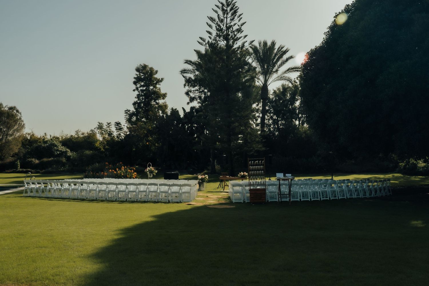 Outdoor wedding ceremony venue with rows of white folding chairs arranged on a lush green lawn surrounded by tall trees.