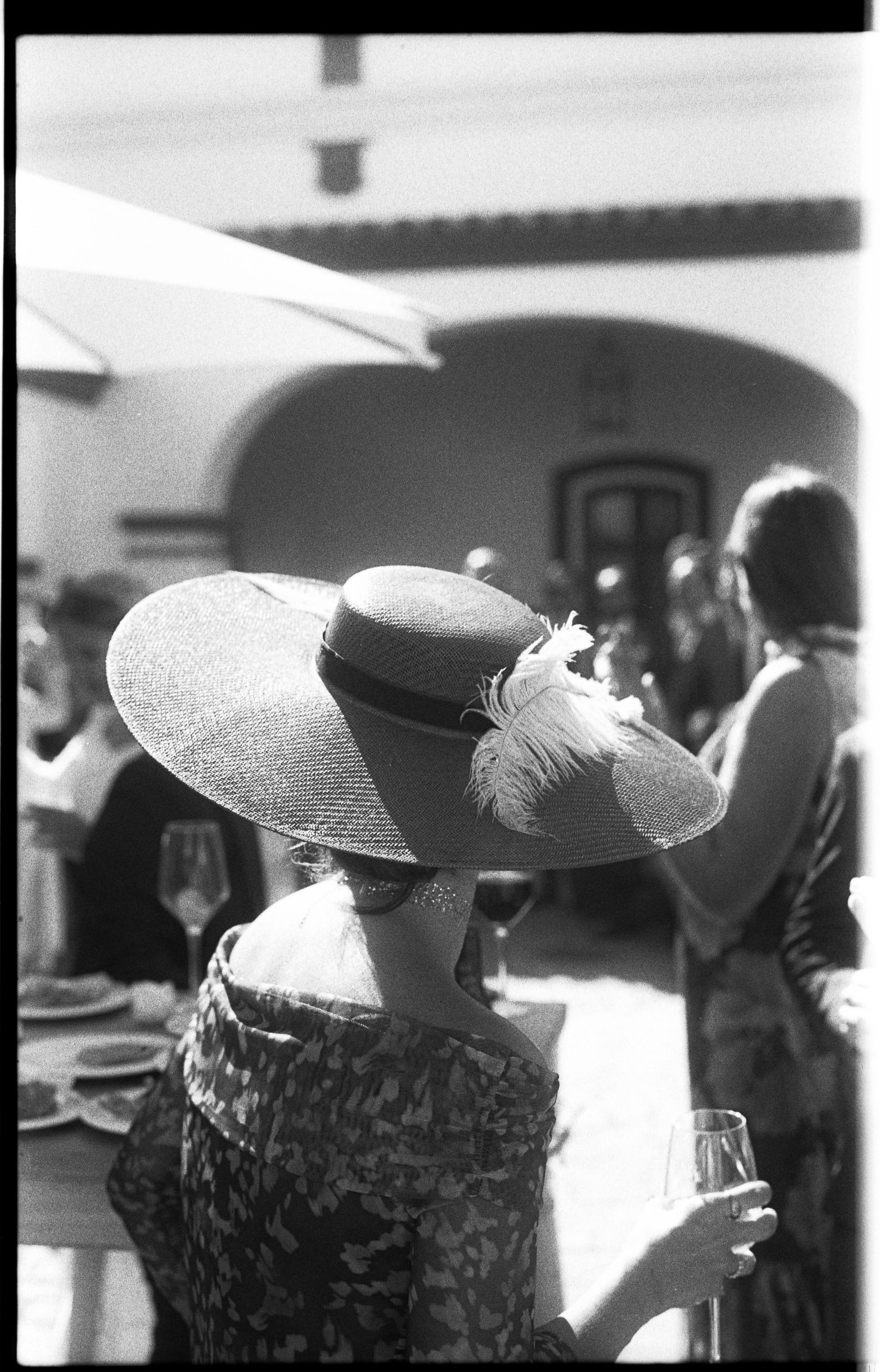Woman wearing wide-brim straw hat with feathers at outdoor wedding reception, black and white film photo.