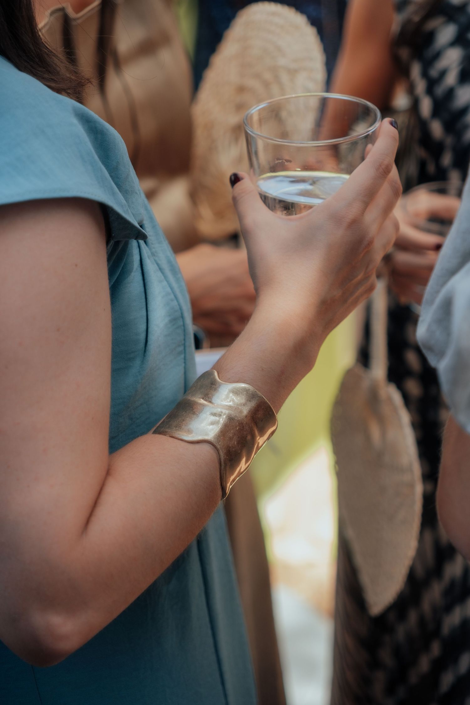 Wedding guest in light blue dress holding a glass of water with a wide gold cuff bracelet accessory.