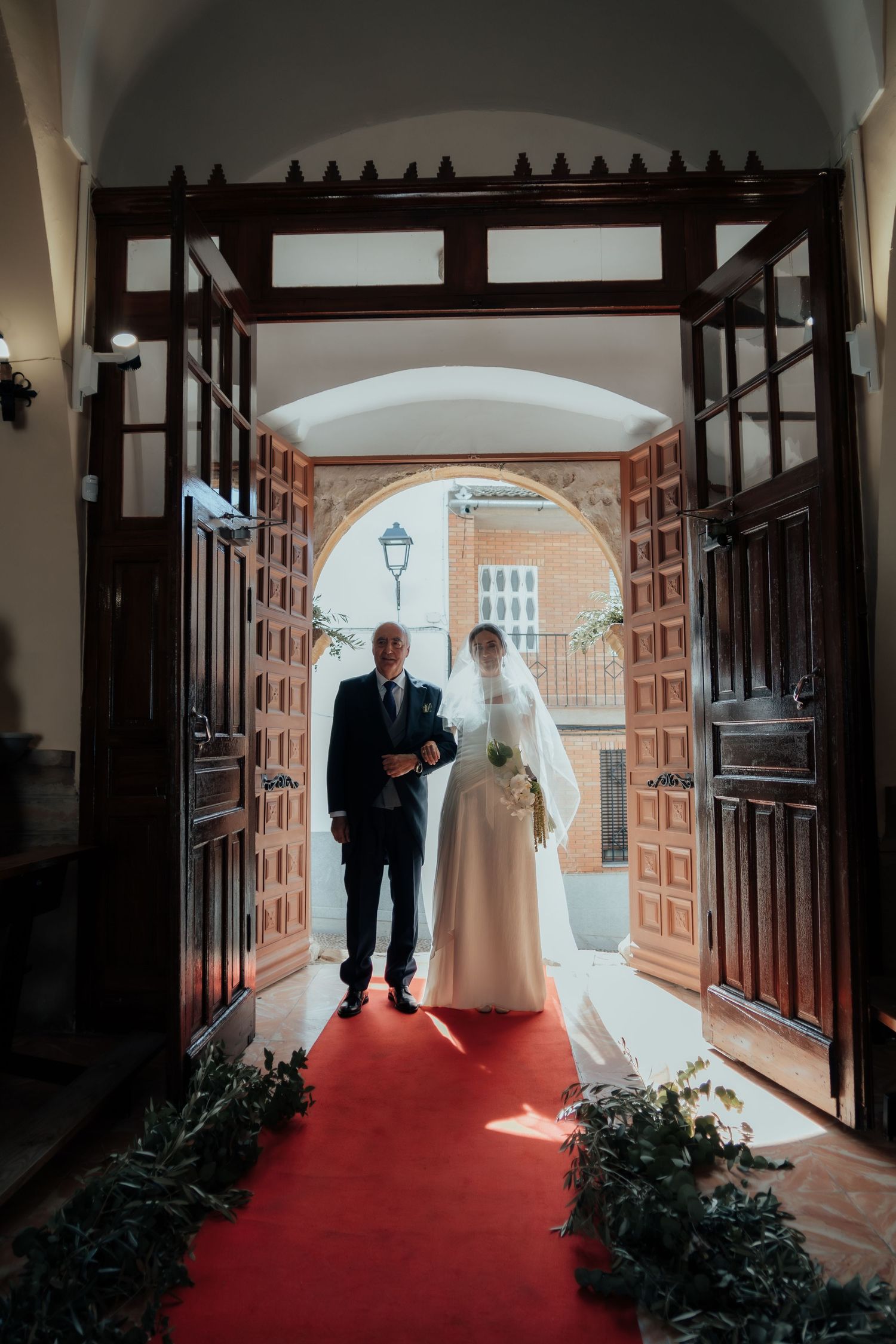 Bride and escort stand at church entrance on red carpet framed by ornate wooden doors.