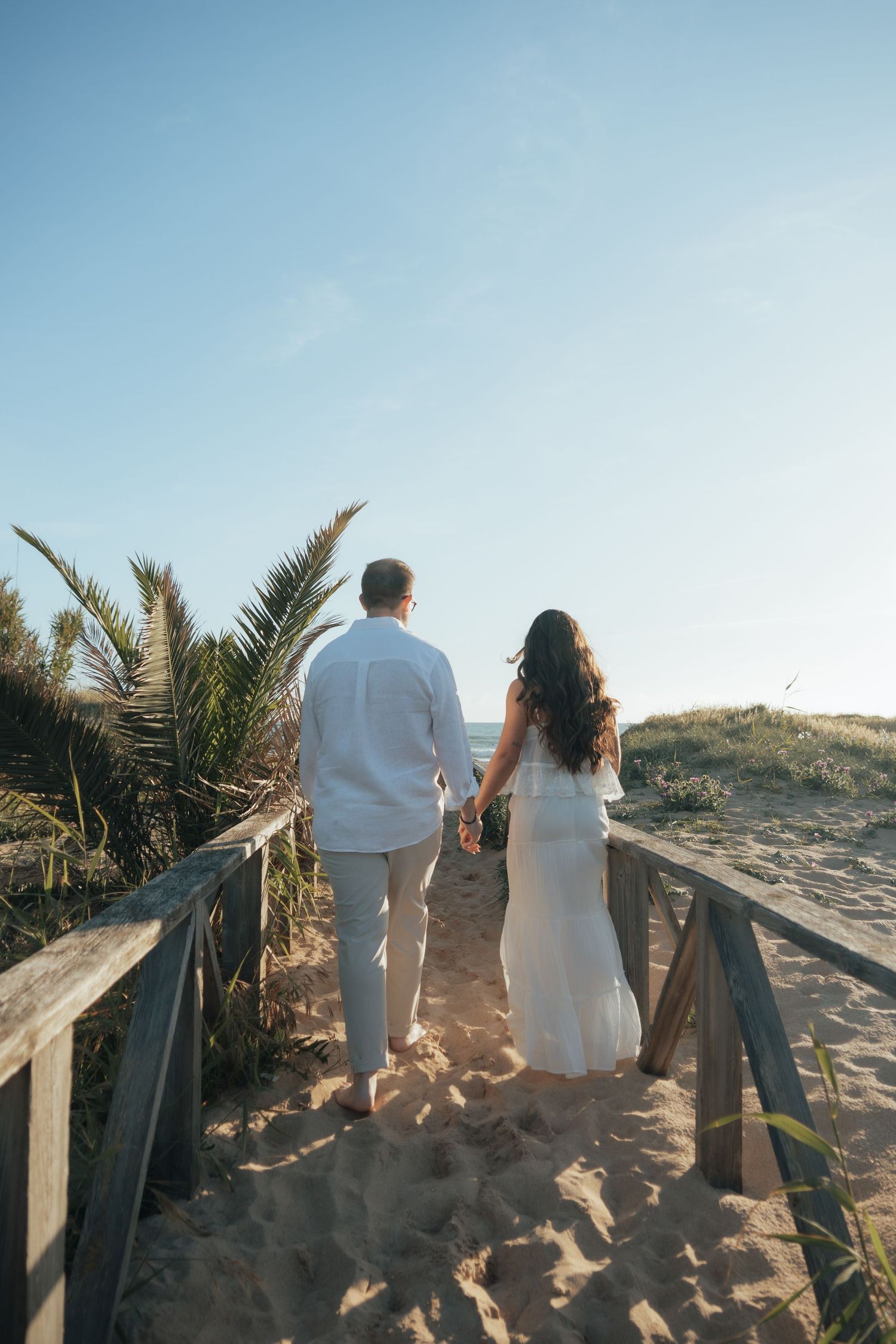 Couple holding hands walking on sandy beach boardwalk path toward ocean, wearing white outfits on sunny day.