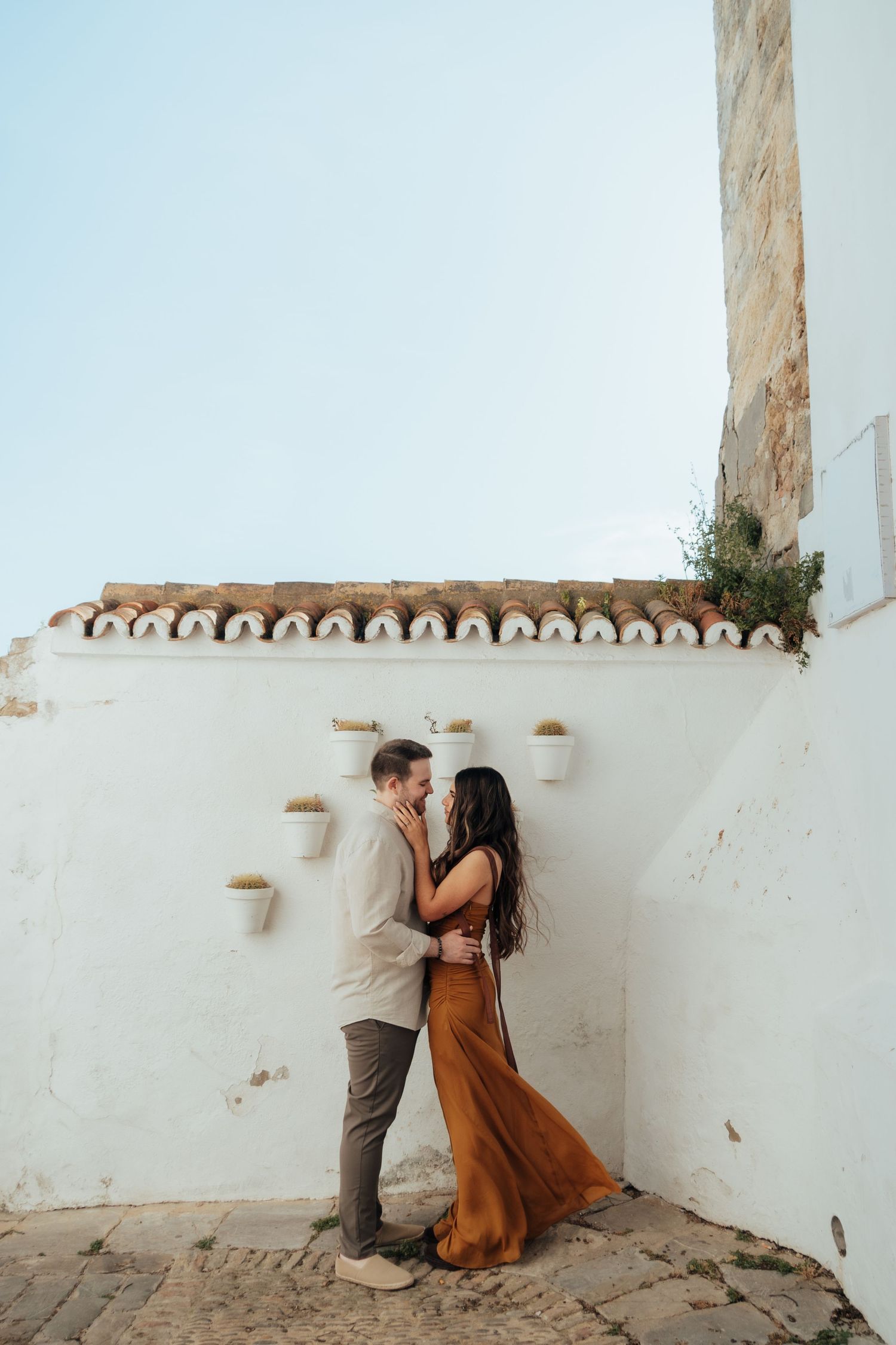 Romantic couple embracing against a white Mediterranean wall with terracotta roof tiles and hanging pots.