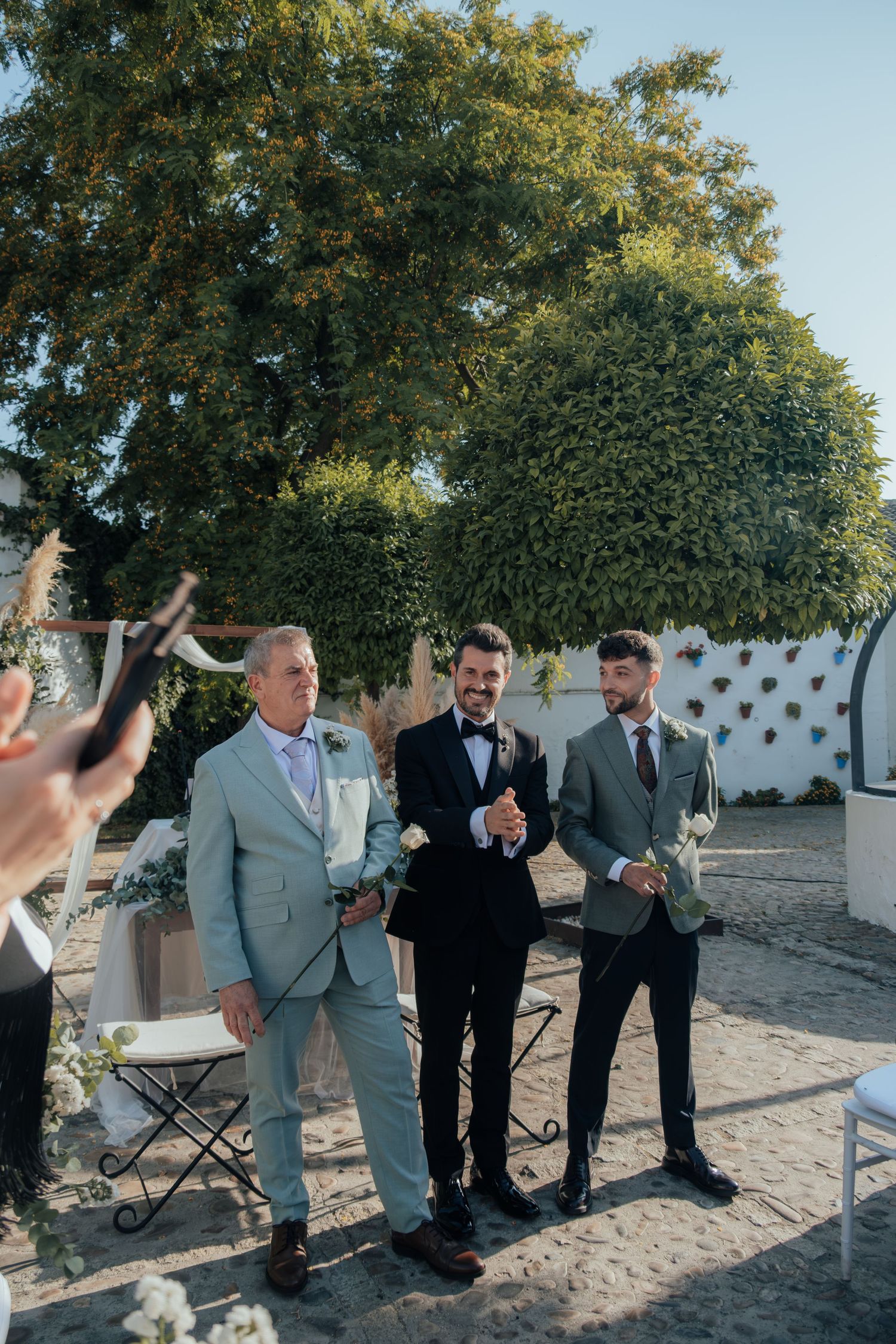 Three men in suits smile and laugh together during an outdoor wedding ceremony celebration.