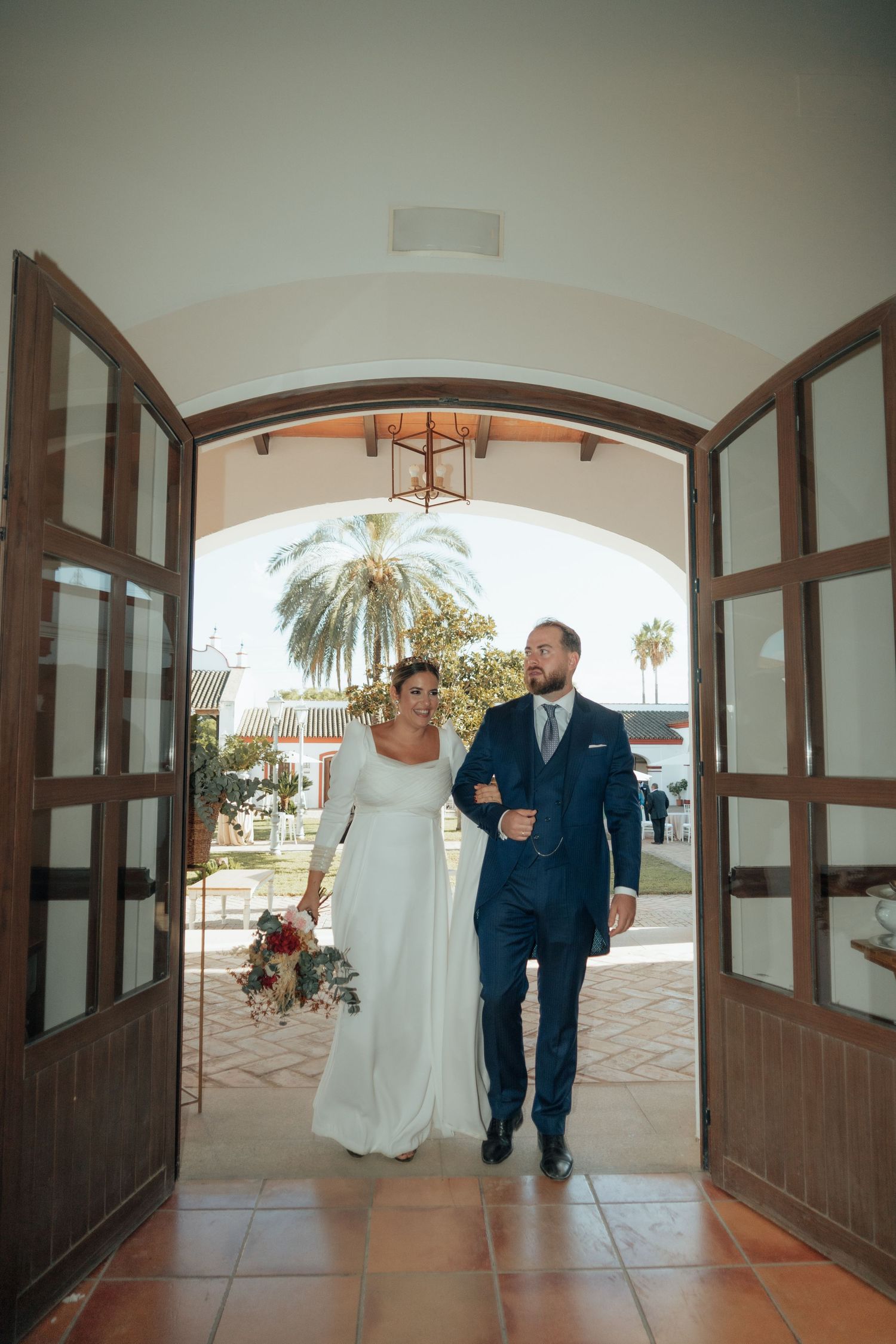 Bride and groom entering through arched wooden doors holding a bridal bouquet of red flowers.