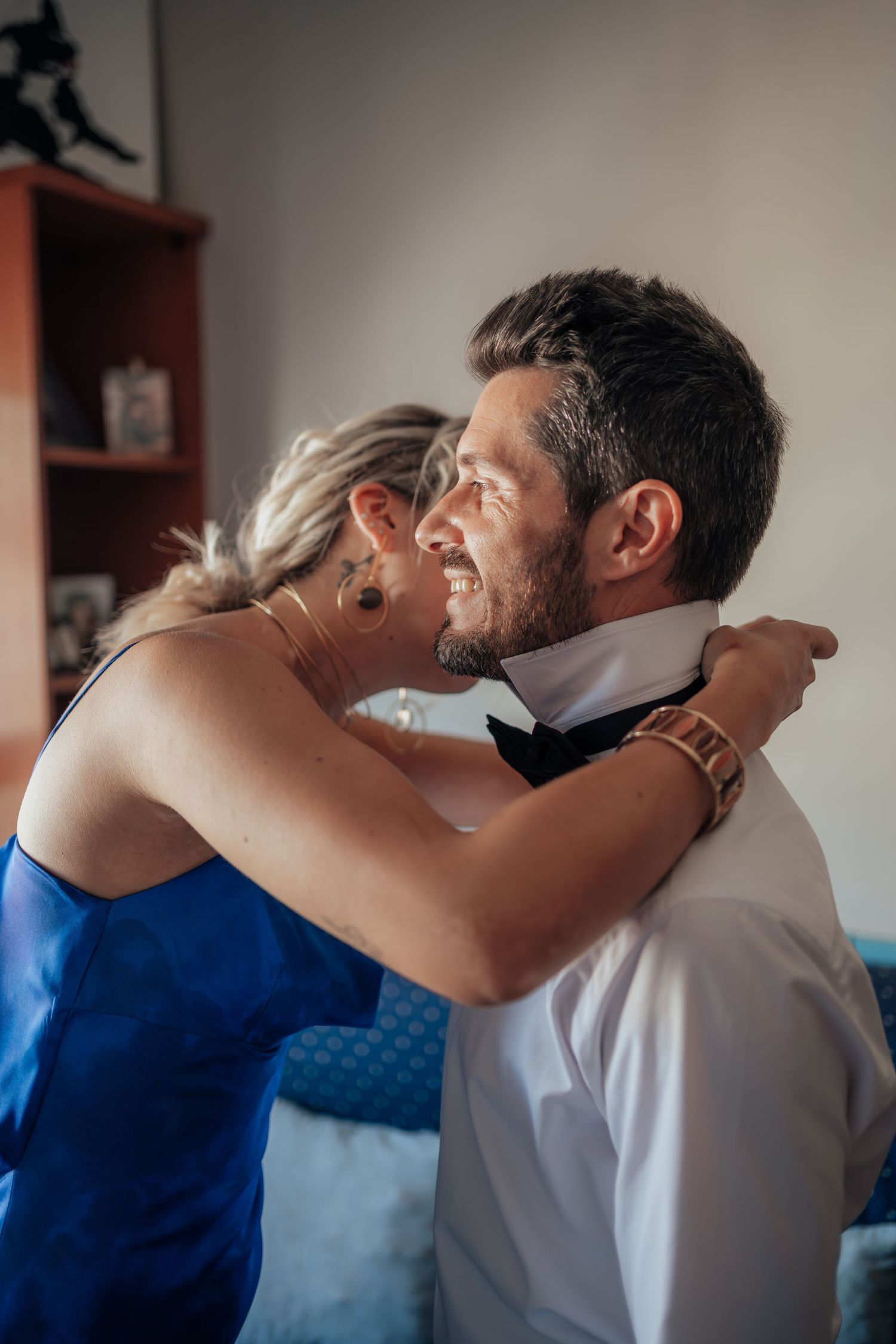 A smiling groom in a white shirt embraces a woman in a blue dress who adjusts his black bow tie.