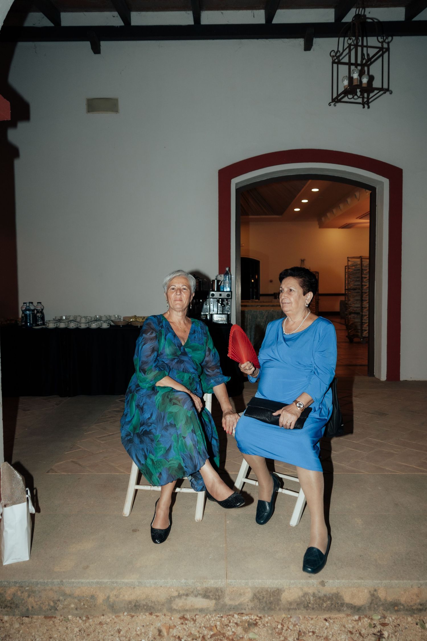 Two elderly women in formal dresses seated on white chairs at an indoor evening event.