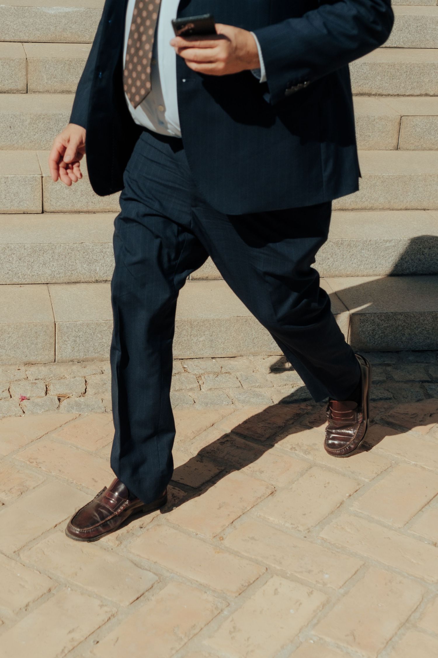 Businessman in navy suit walking down stone steps while using smartphone outdoors.