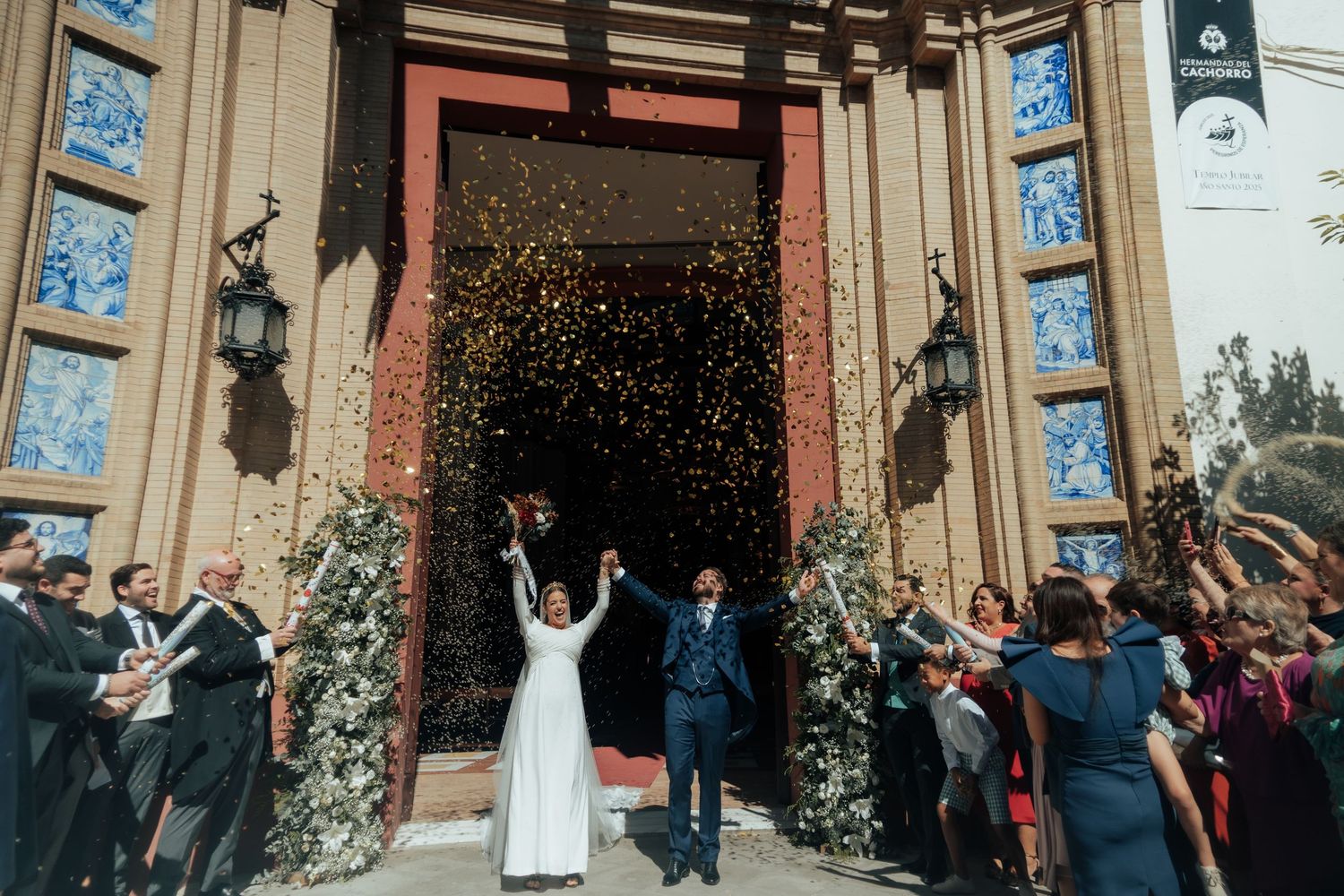 Bride and groom celebrate with raised arms as colorful confetti fills the air outside church.