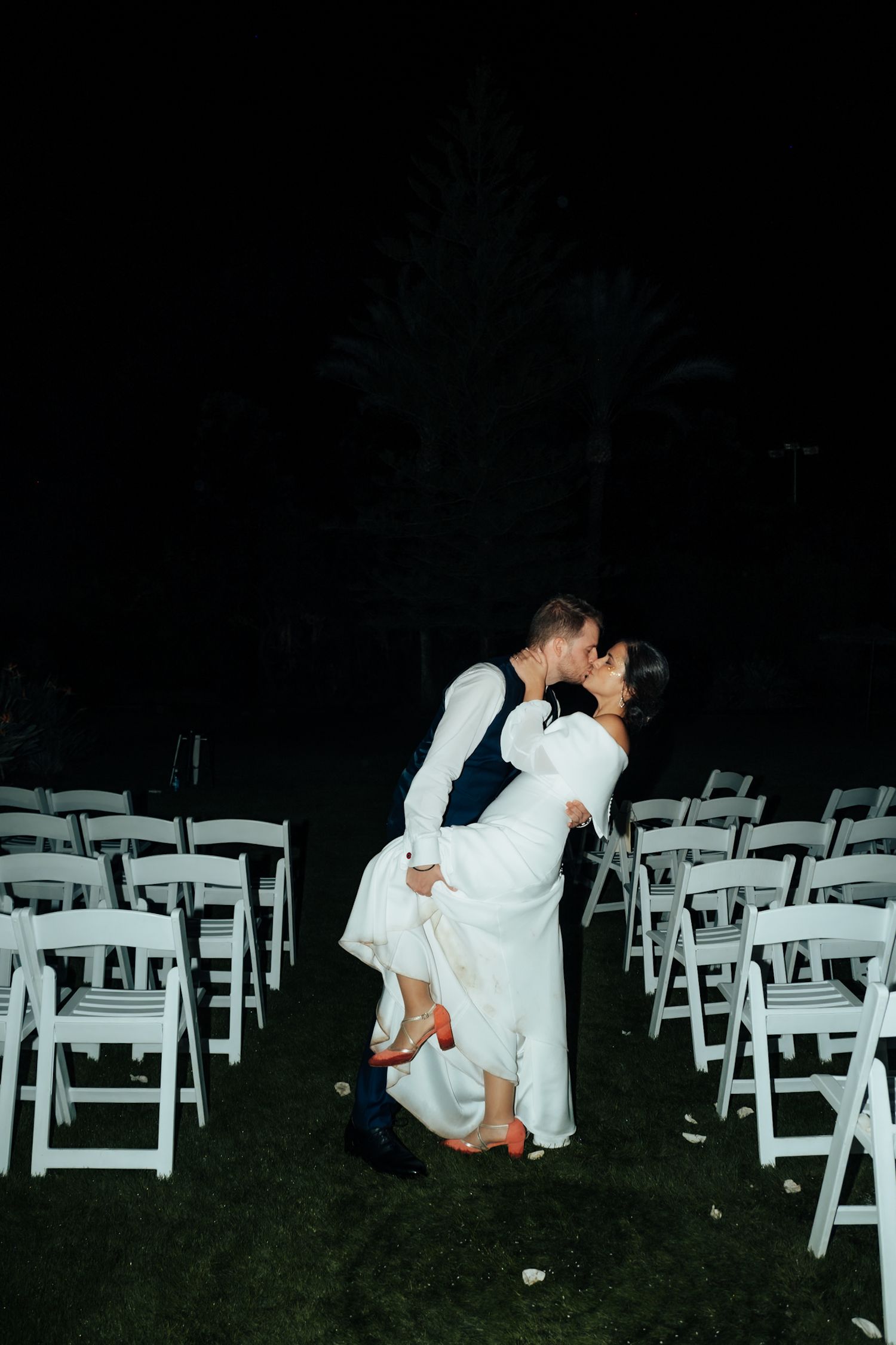 Groom dips bride in romantic kiss between rows of white chairs at dark outdoor wedding ceremony venue.