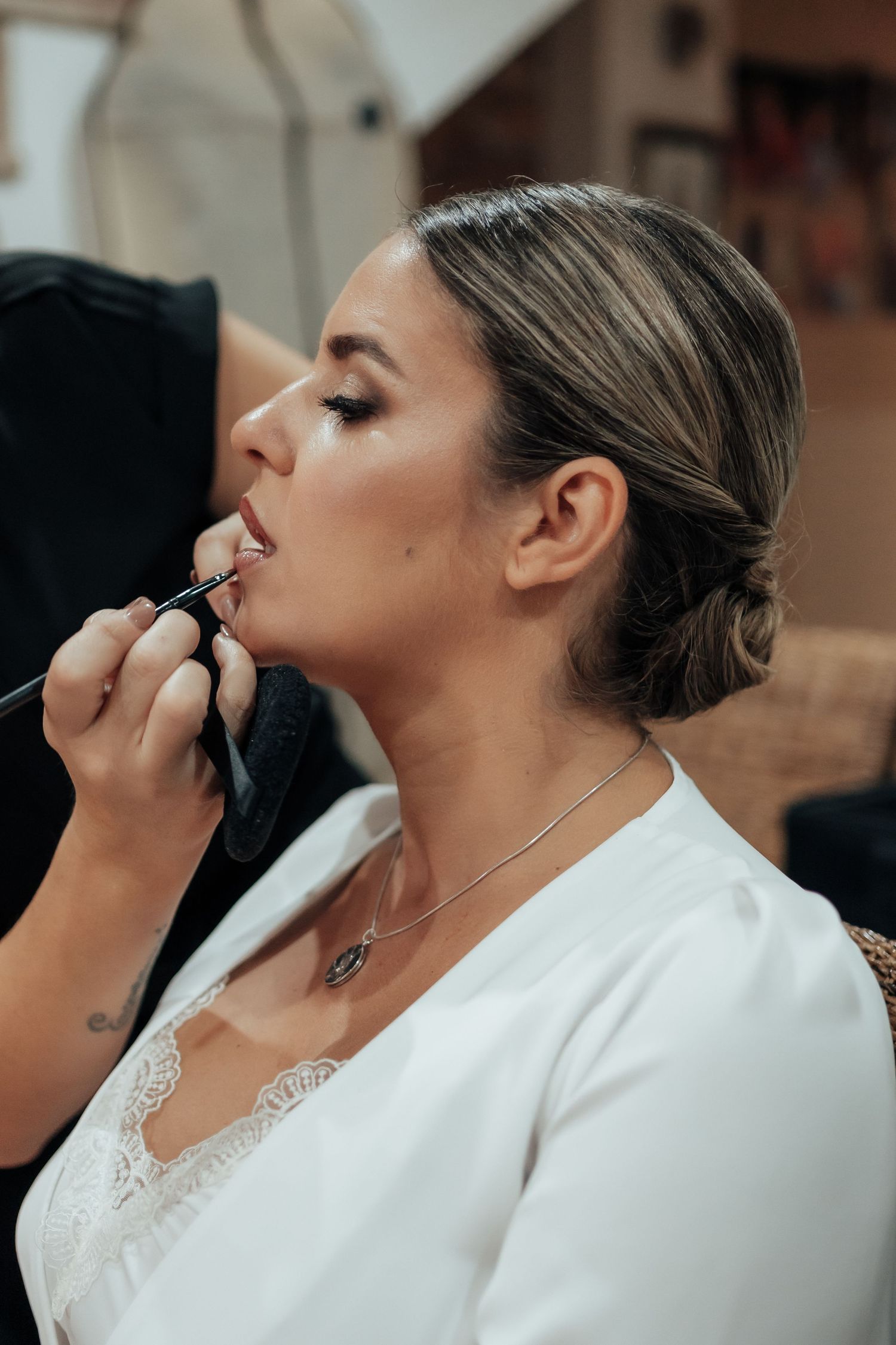 Makeup artist applying lipstick to a bride with an elegant updo wearing a white lace robe.
