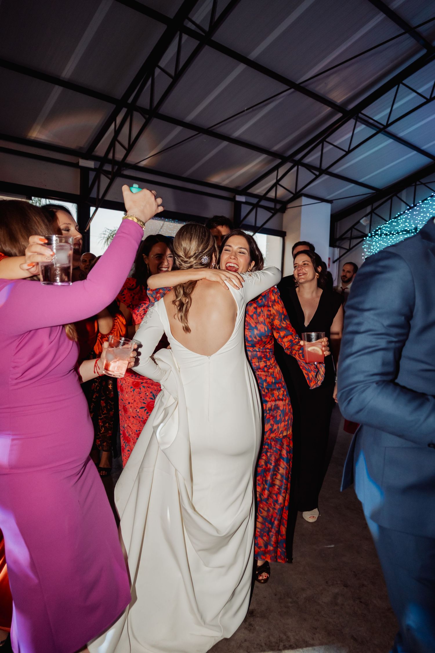 Bride in backless white gown receiving warm hugs from guests at a lively wedding reception celebration.