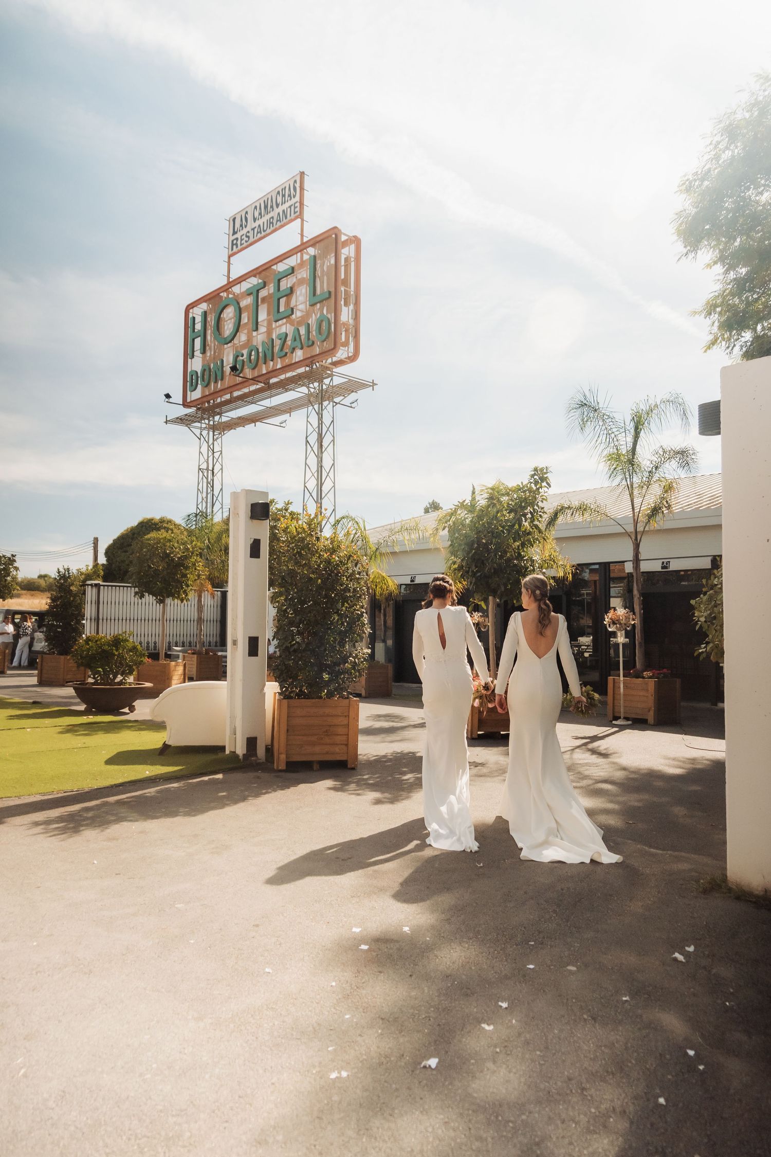 Two brides in white gowns walk hand in hand toward Hotel Don Gonzalo on sunny day.