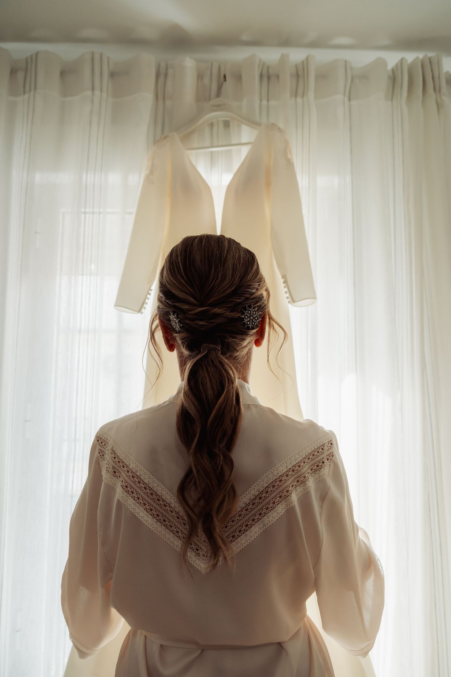 Bride with braided updo gazing at her ivory wedding dress hanging by sheer curtains.