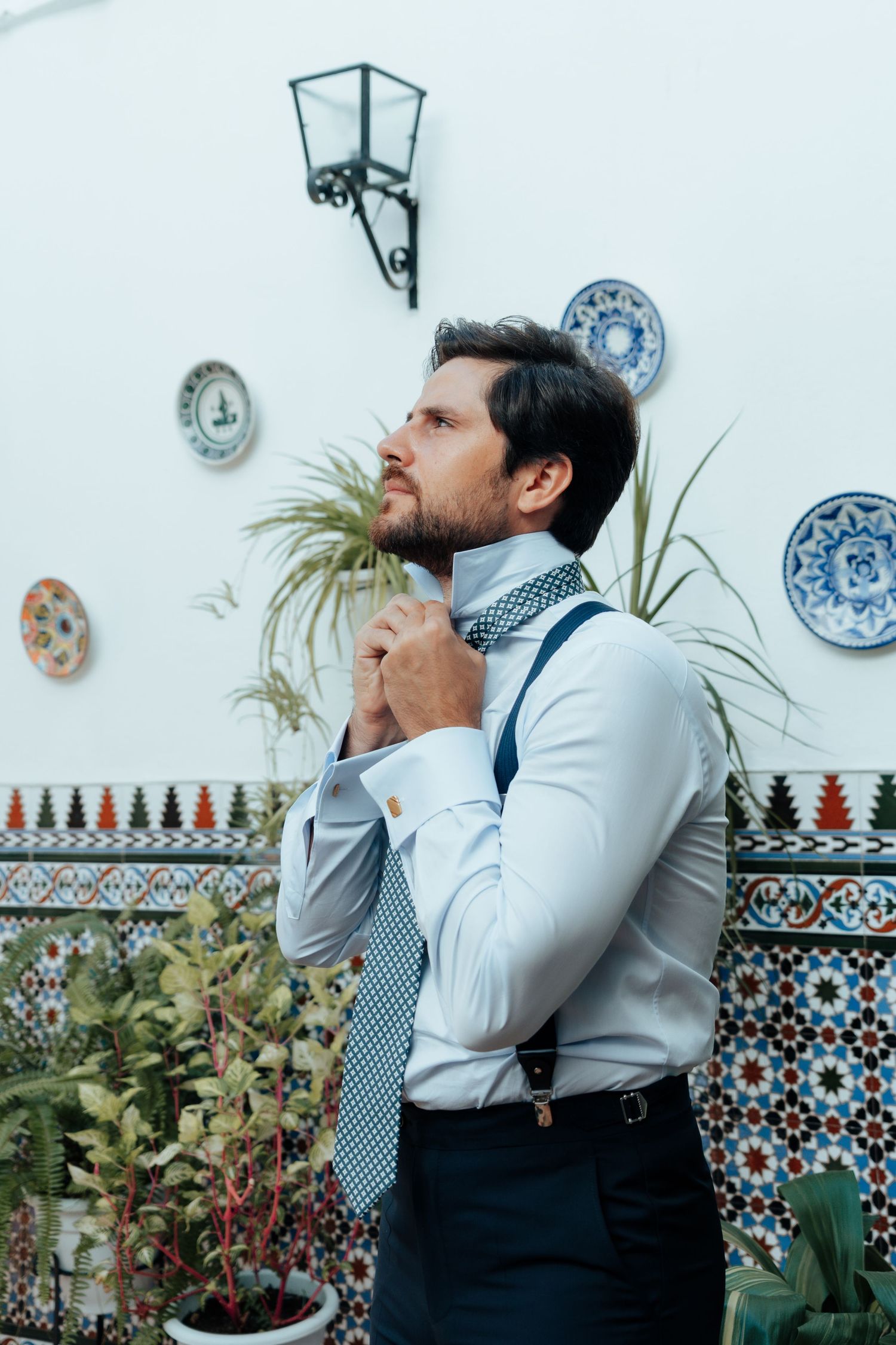 Stylish groom adjusting patterned tie wearing suspenders in Spanish tiled courtyard with plants.
