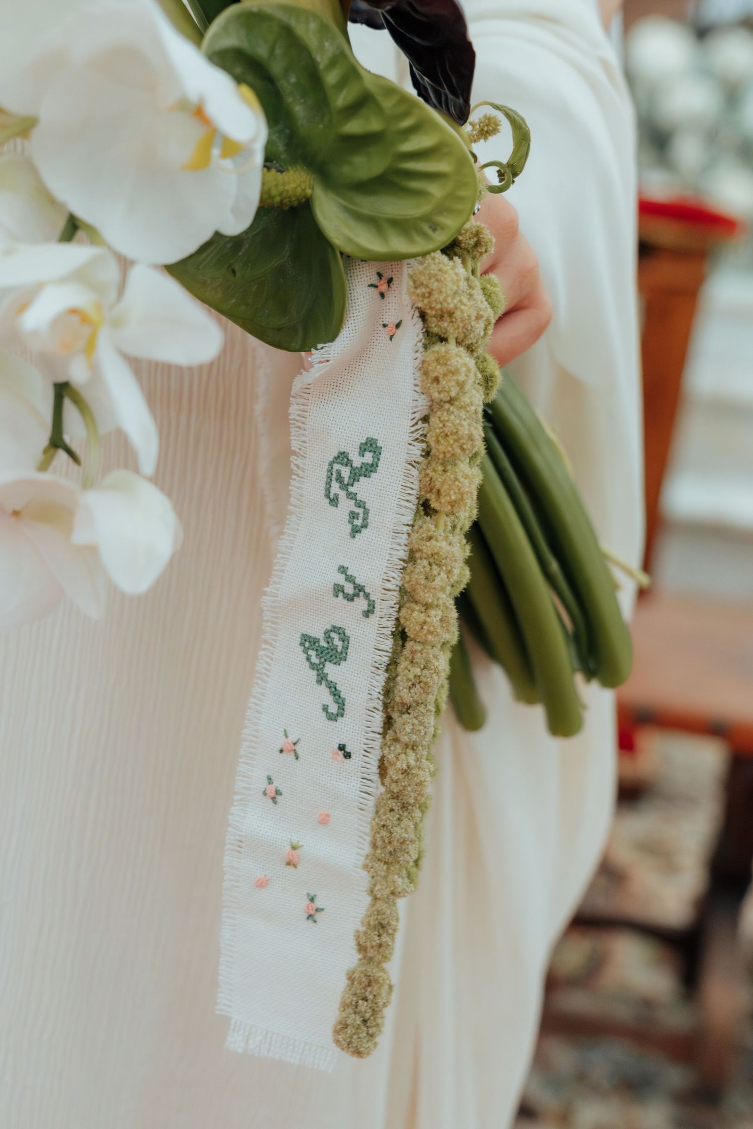 Close-up of bridal bouquet with white orchids and embroidered ribbon monogram detail in green lettering.