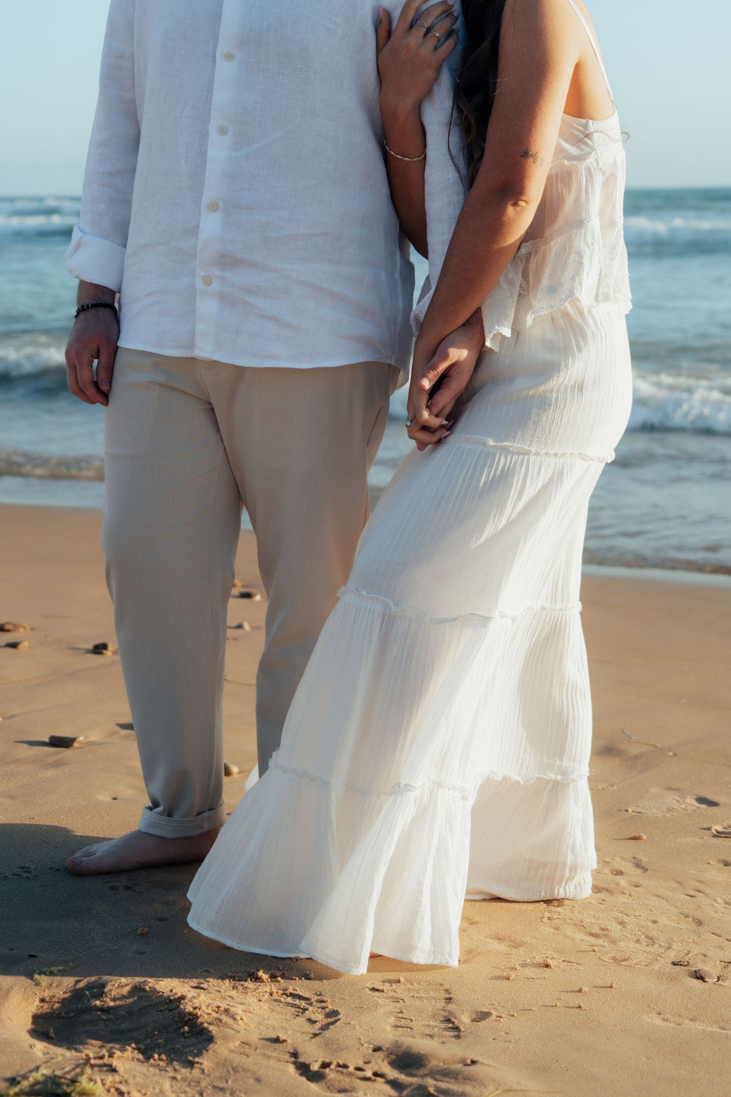 Couple holding hands on sandy beach, woman in white tiered dress, man in linen shirt and khaki pants, ocean waves behind.