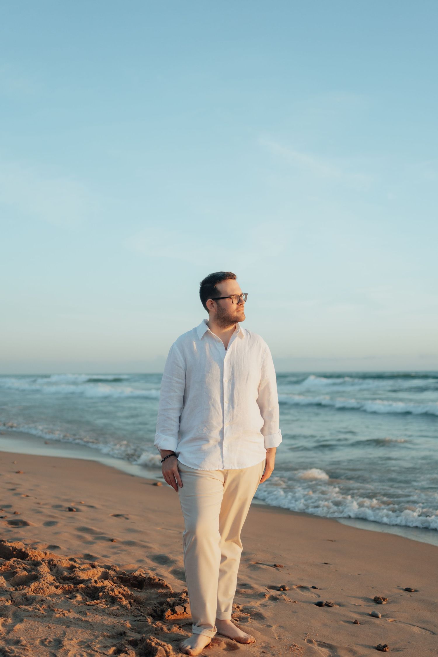 A man in a white linen shirt and beige pants walks barefoot on a sandy beach at sunset.