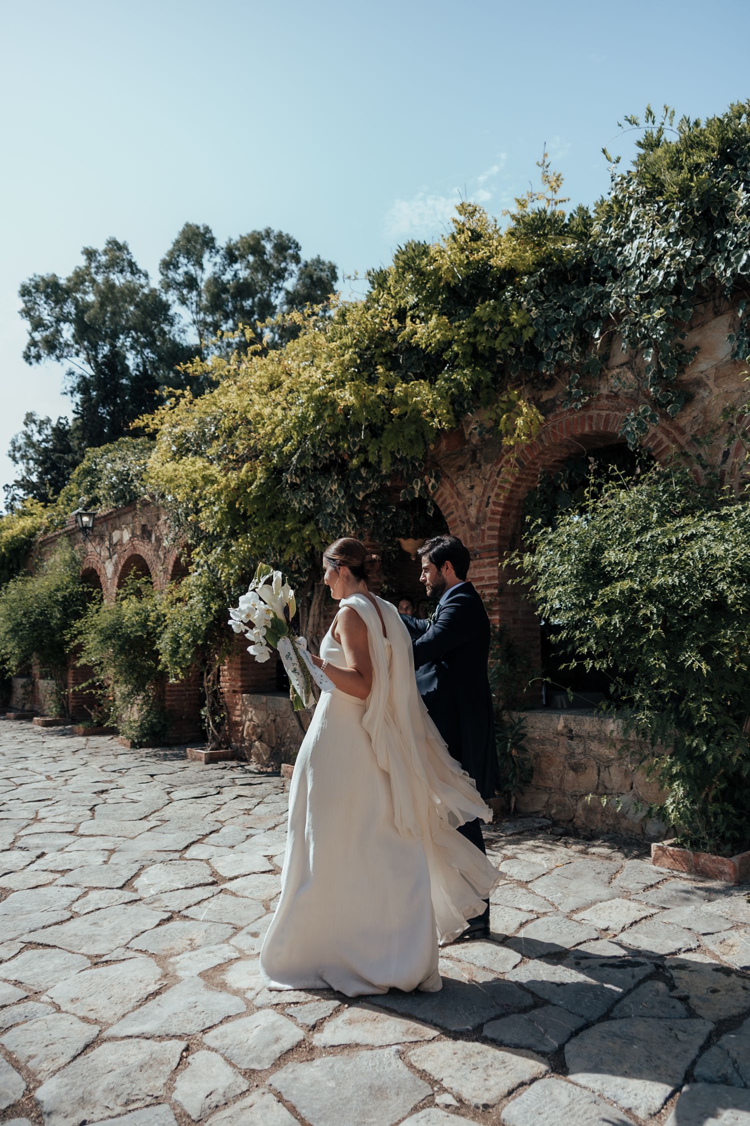 Bride and groom walking along a stone courtyard beside a brick arch wall covered in lush green foliage.
