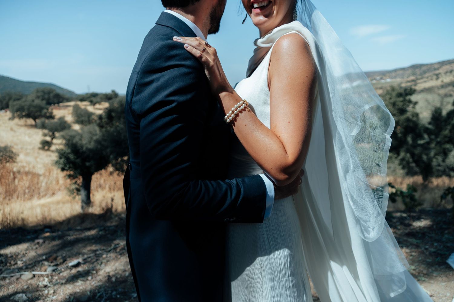 Close-up of bride in white gown with veil and groom in navy suit embracing in sunny countryside.