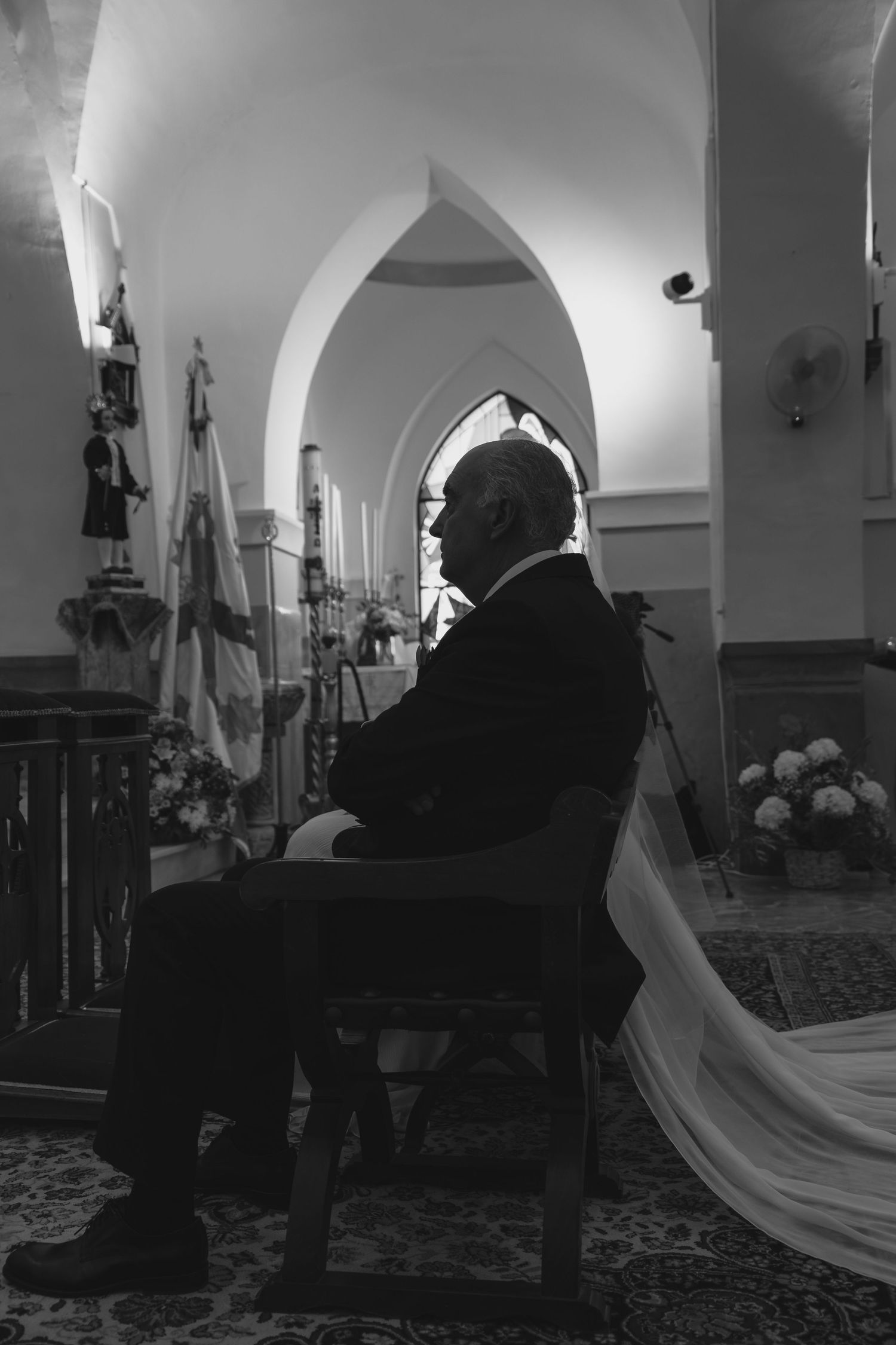 Black and white photo of elderly man seated in church during wedding ceremony with bridal veil.
