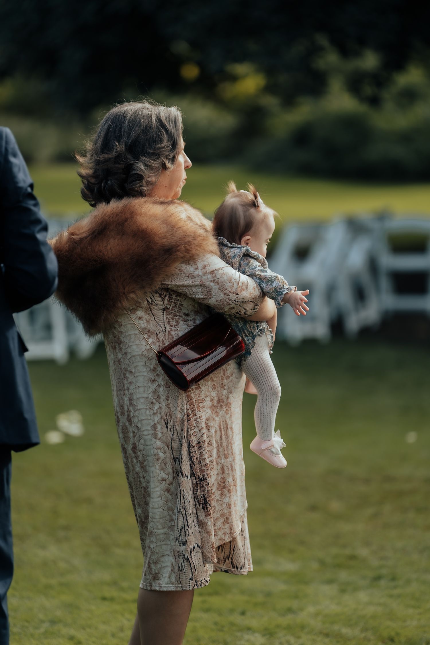 Elegant grandmother in fur stole and floral dress tenderly holds a baby at an outdoor wedding ceremony.
