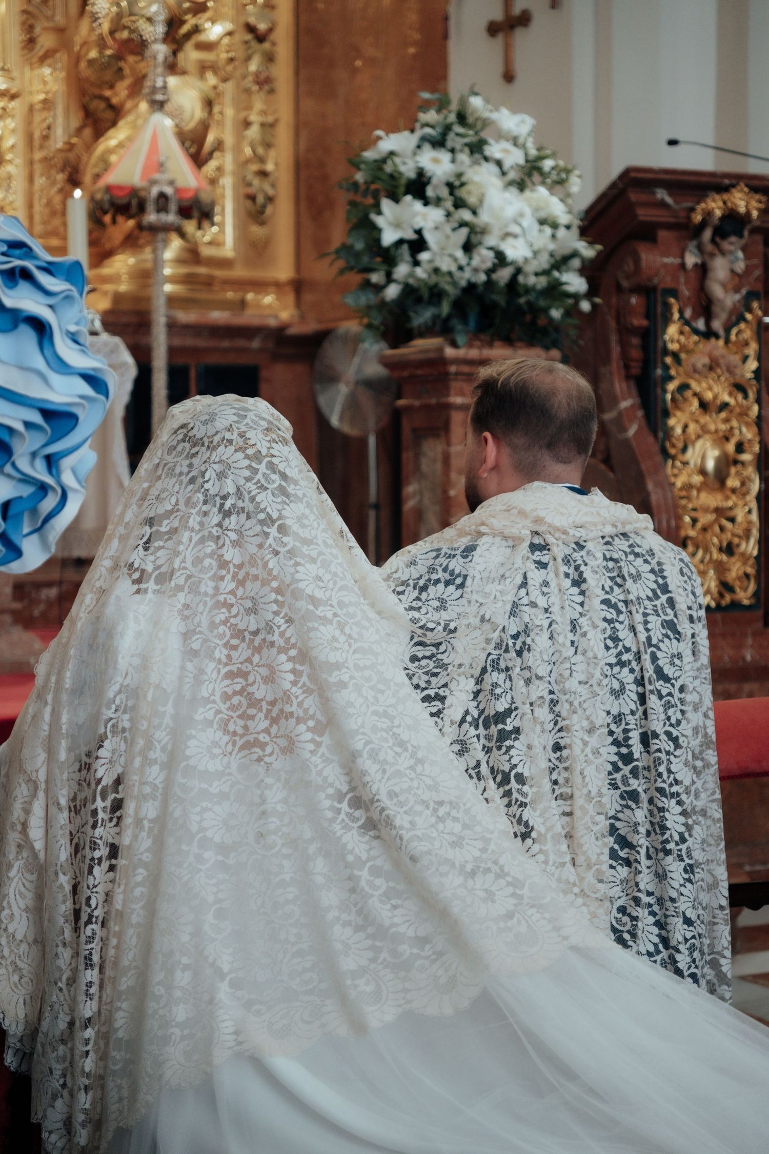 Bride and groom kneeling at church altar wearing traditional Spanish lace mantilla during wedding.