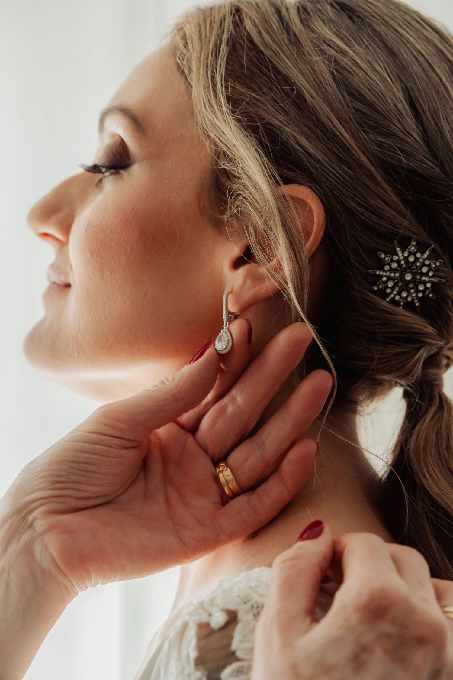 Bride in profile smiling as hands help place elegant crystal drop earring near her ear.