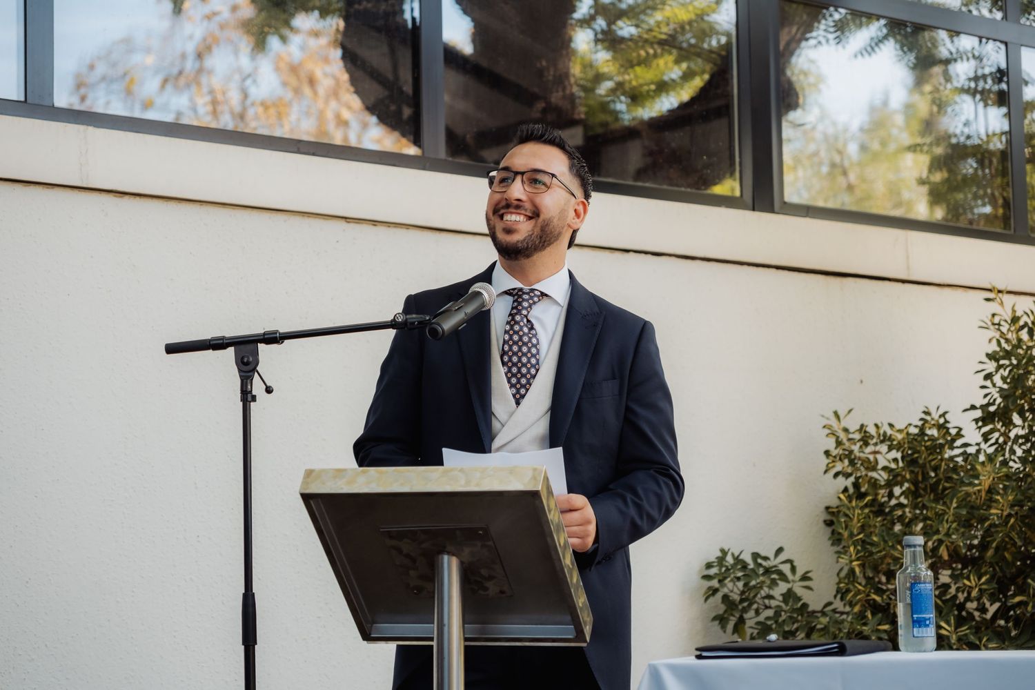 Smiling man in navy suit and tie speaks at podium with microphone at outdoor wedding ceremony.