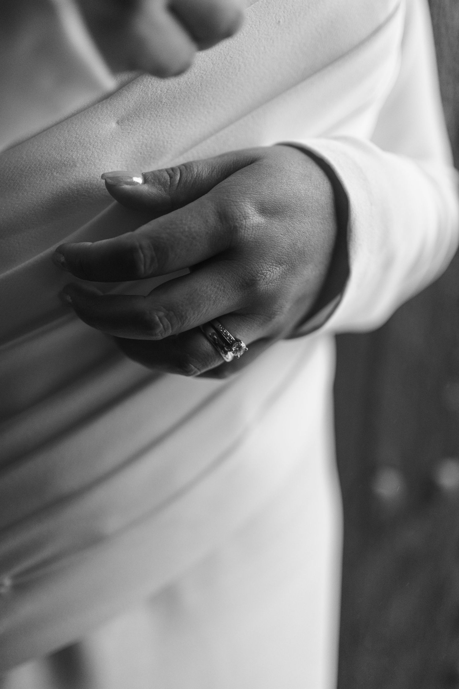 Black and white close-up of a bride's hand wearing an elegant engagement ring on her white wedding dress.