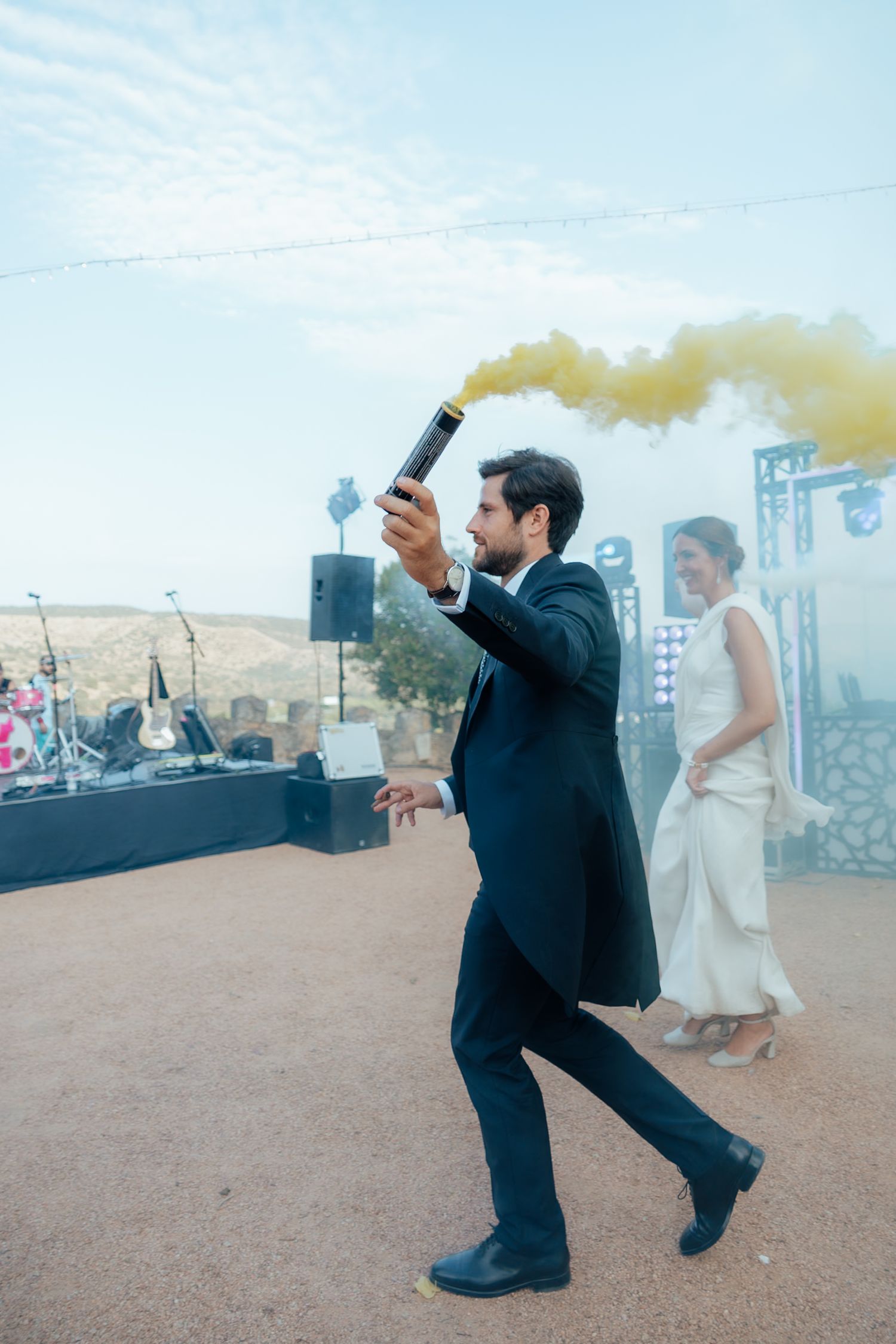 Groom in black suit releasing yellow smoke flare at outdoor wedding celebration entrance.